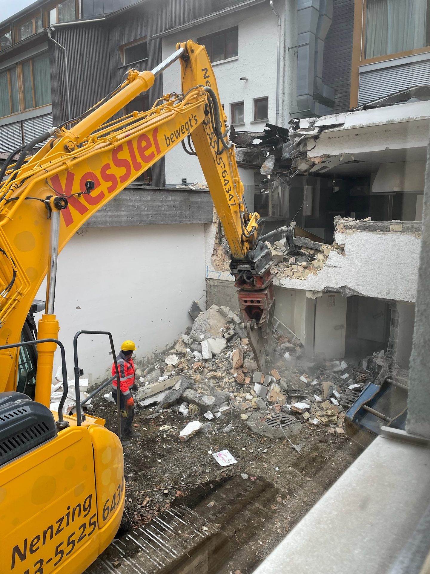 An excavator demolishes a building, with a worker standing amidst debris on the construction site.