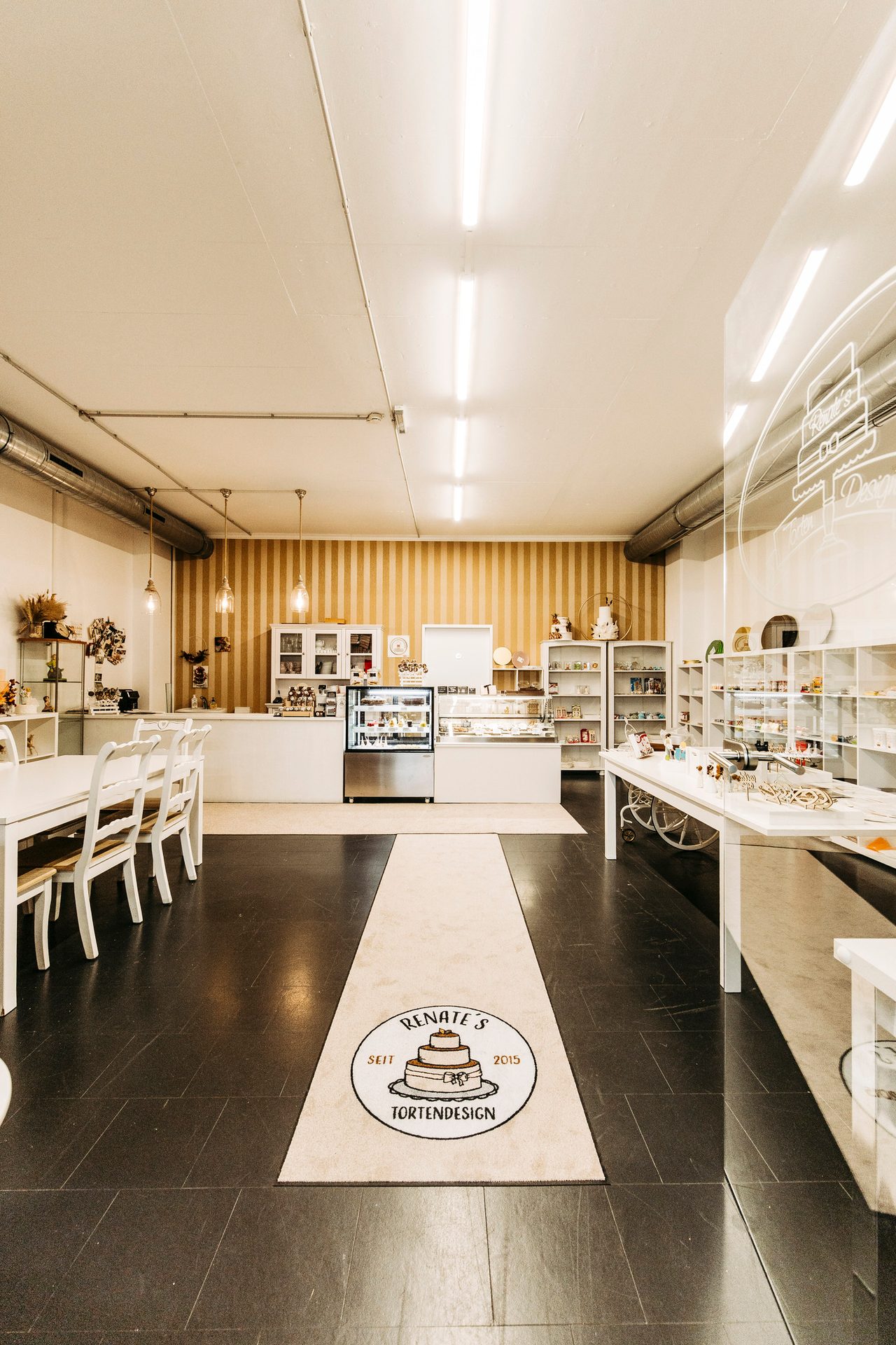 Bright cake shop interior with display cases, customer seating, and a "Renate's Tortendesign" logo rug.