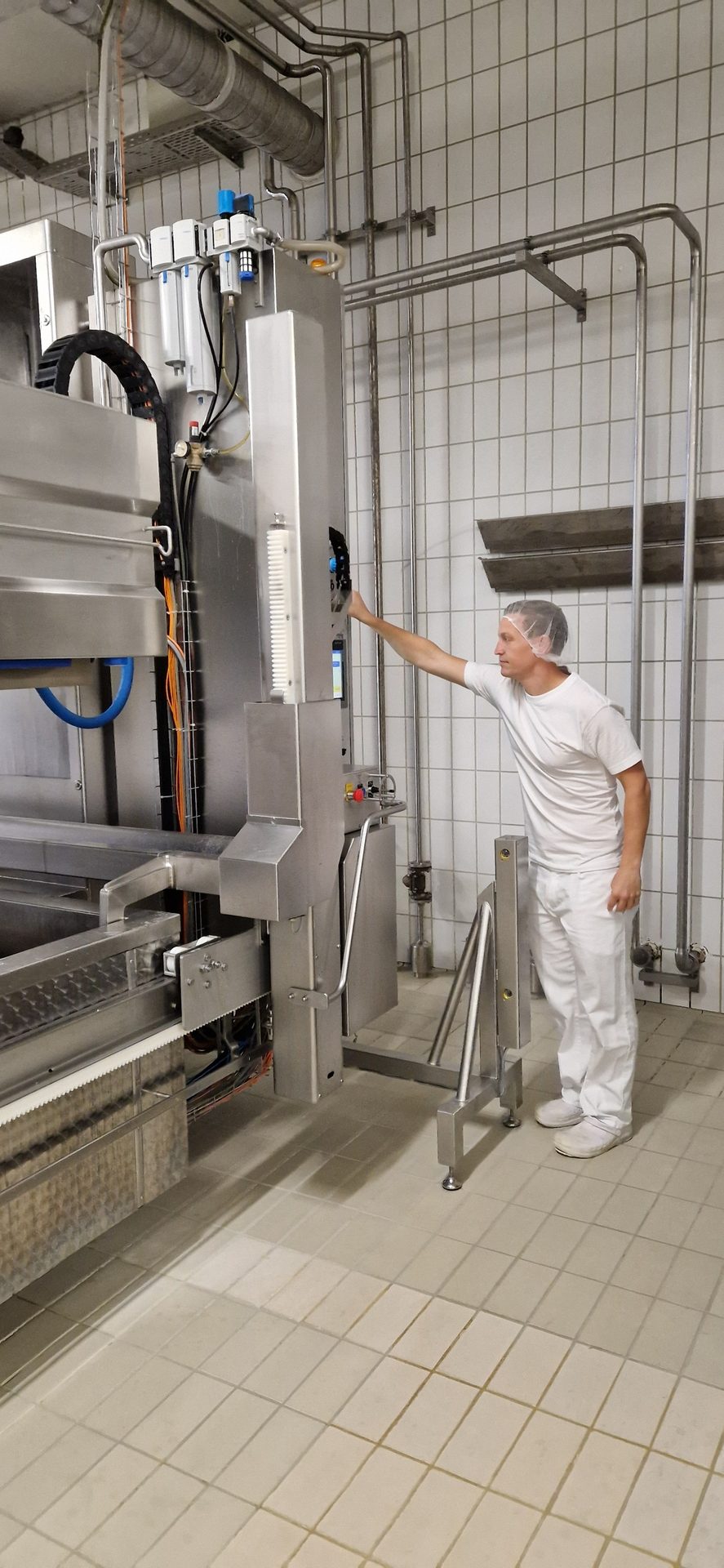 Man in hygienic uniform adjusts stainless steel industrial machinery in a clean, tiled room.
