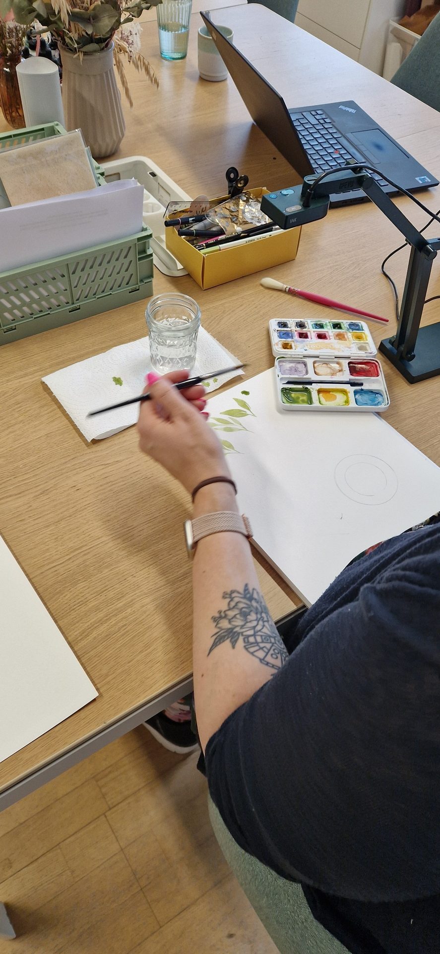 Person painting green leaves with watercolors. Art supplies, laptop, and dried flowers on a table.
