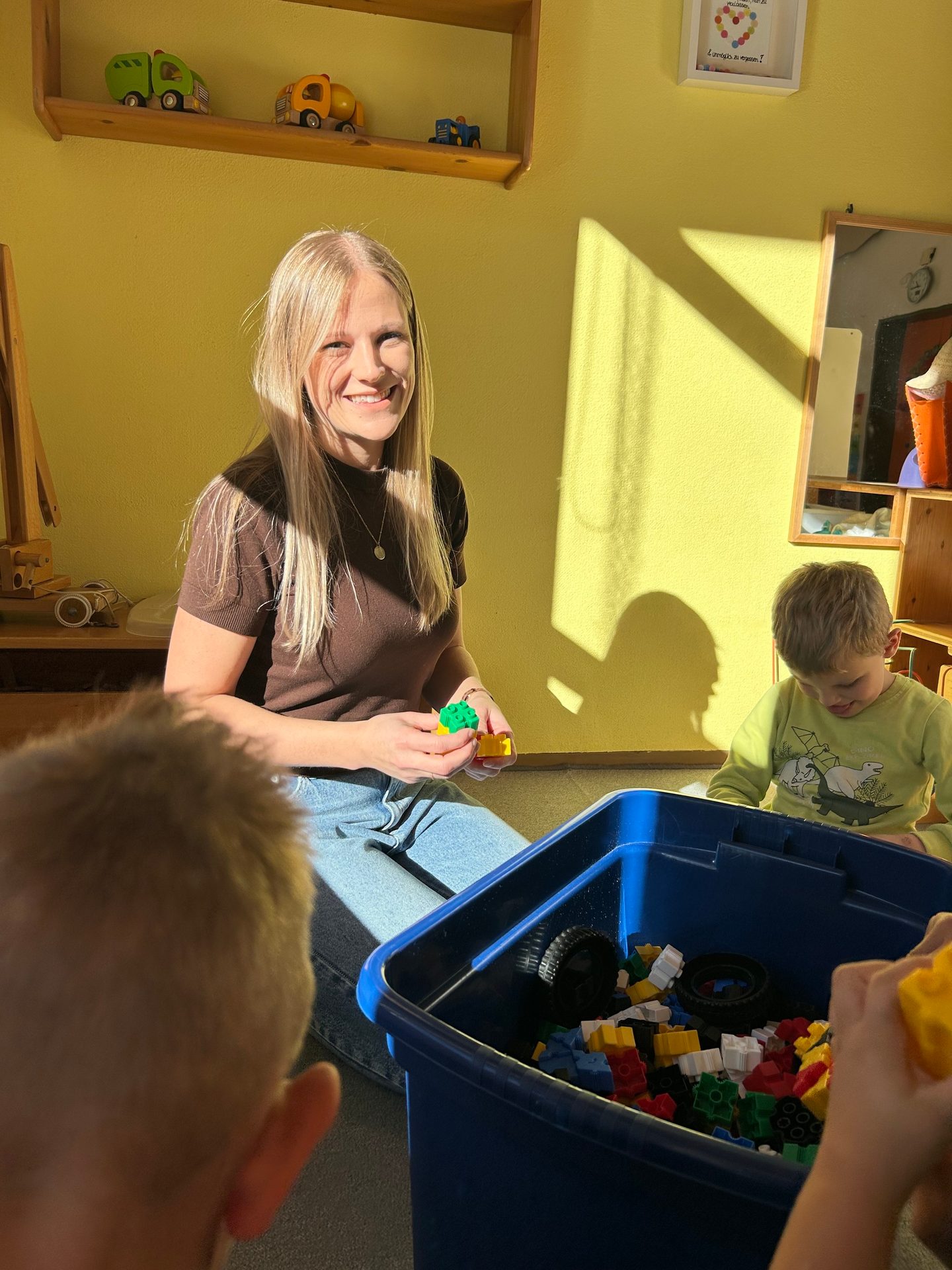 A smiling woman and two children play with building blocks in a sunlit room.