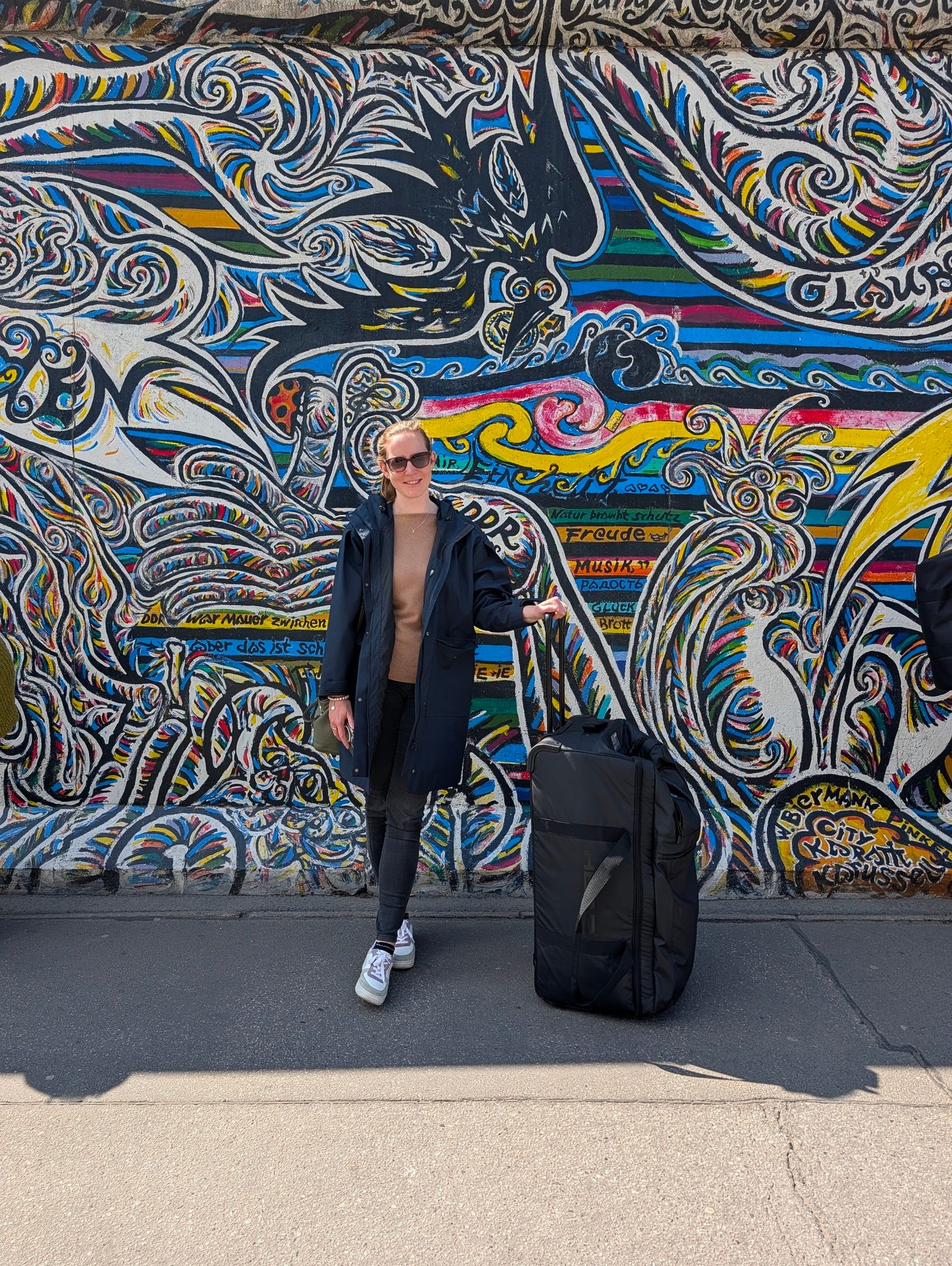 A woman stands next to a large black duffel bag against a vibrant, graffiti-covered section of the Berlin Wall.