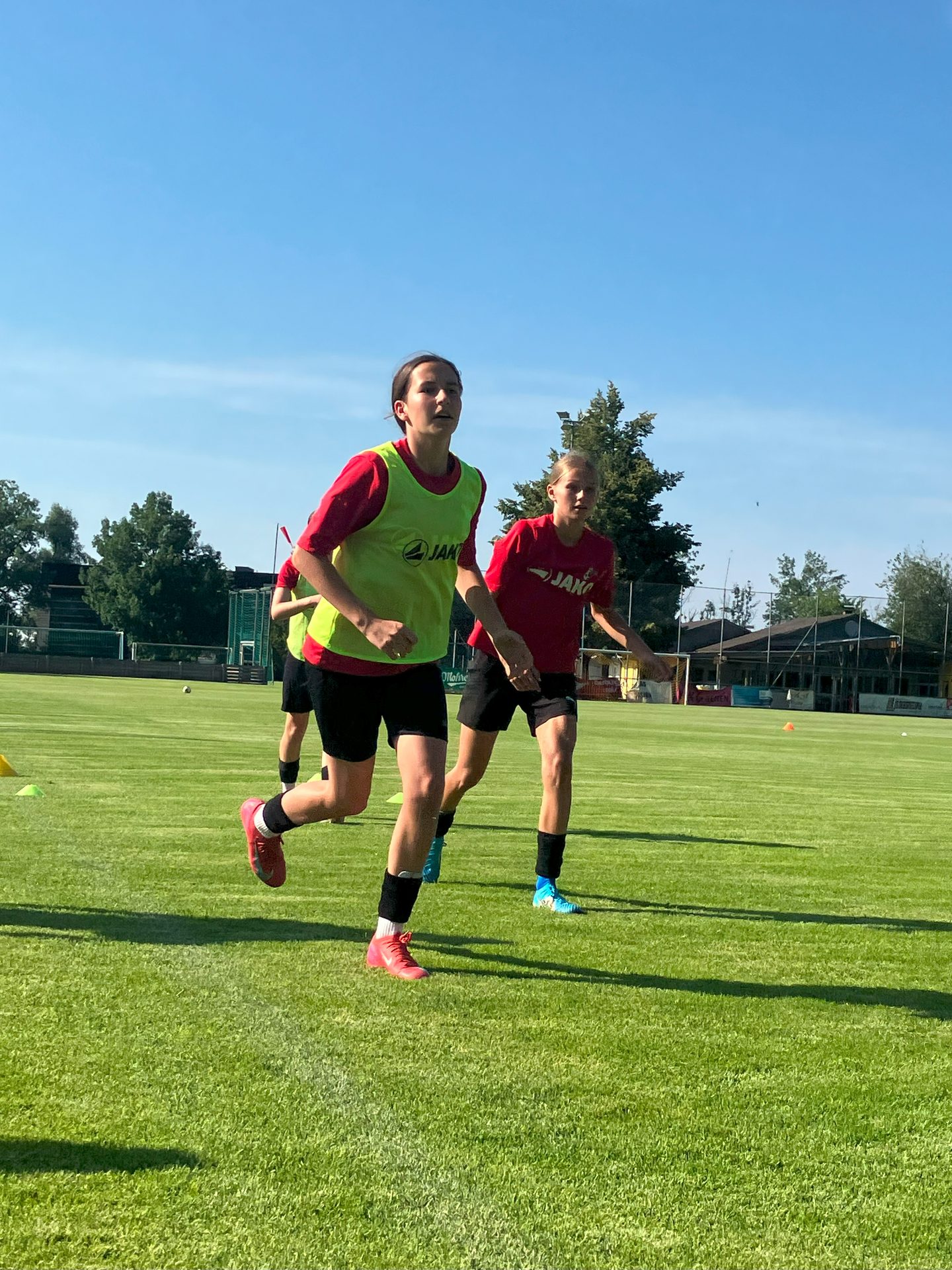 Two female soccer players run on a sunny green field, one in a red shirt and yellow training bib.