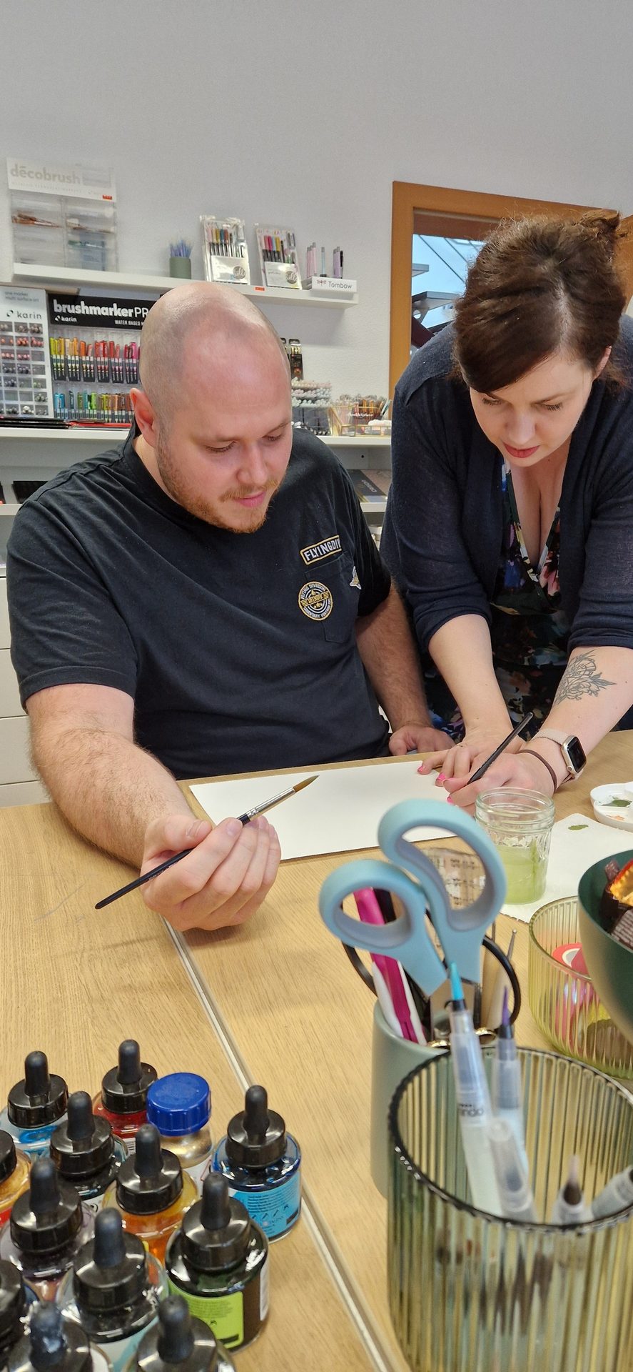 Two people using brushes on paper at a table with art supplies.