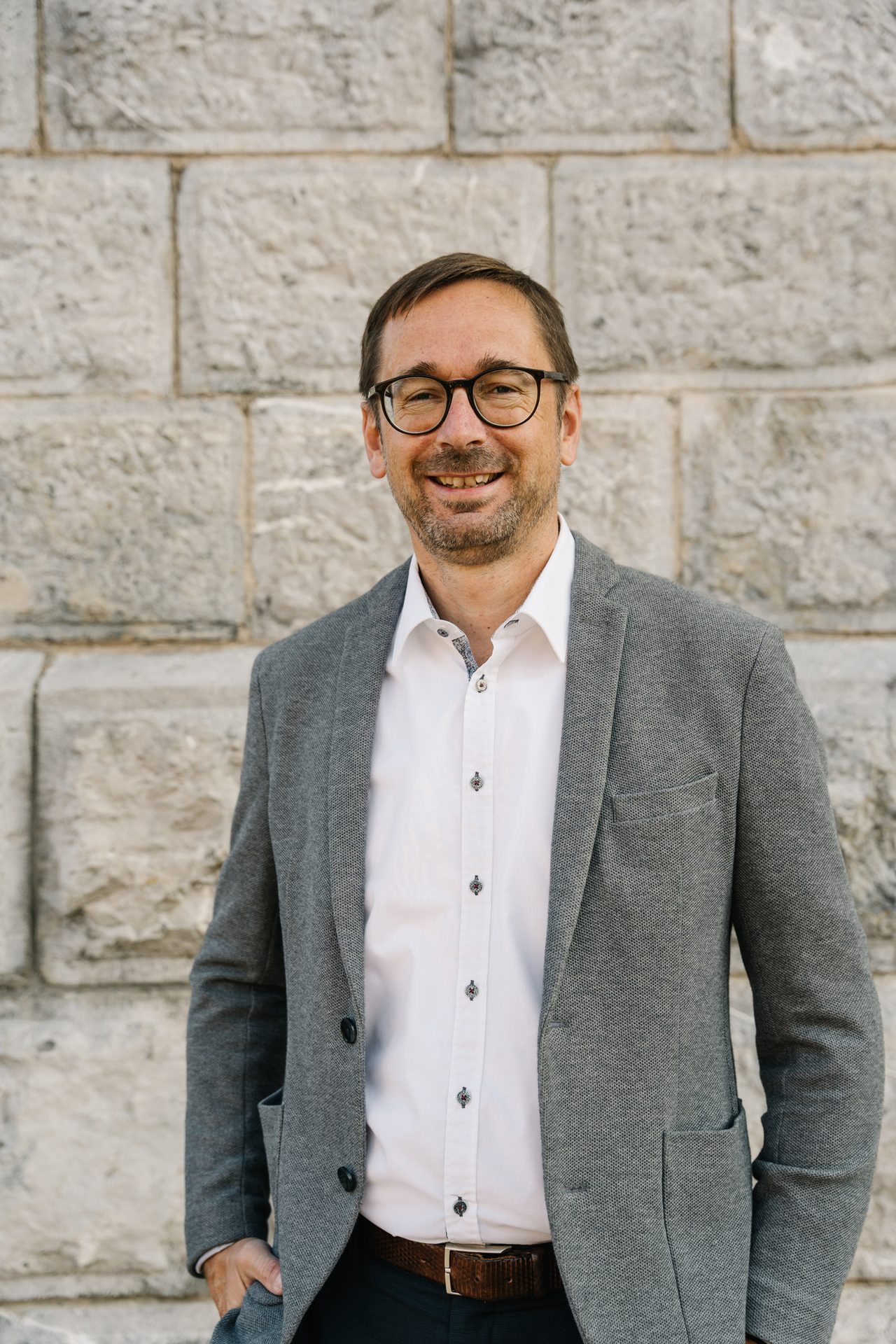 Smiling man in glasses, grey jacket, white shirt, against a stone wall.