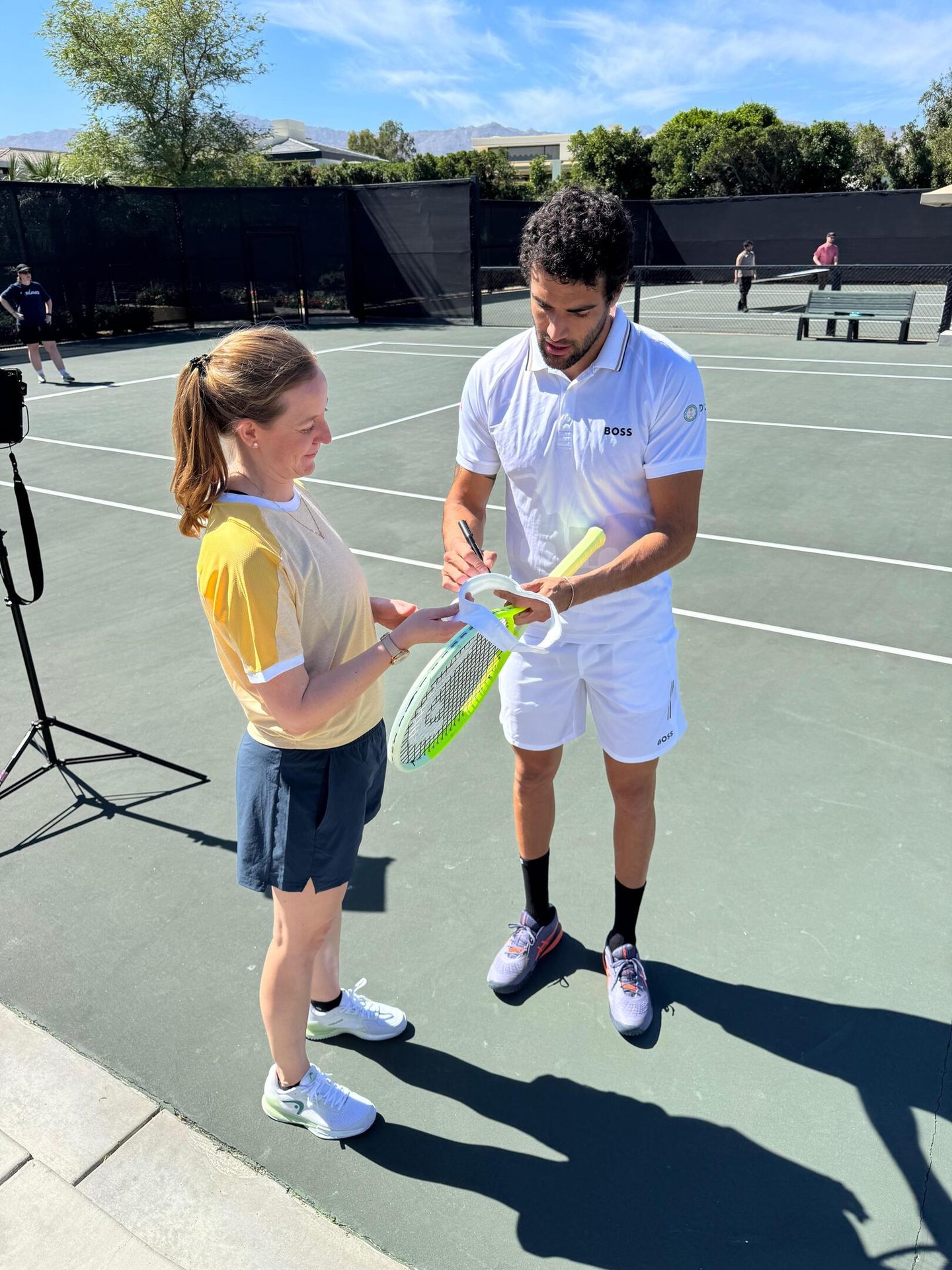 A man in a white polo shirt signs a tennis racket held by a woman in a yellow shirt on a tennis court.