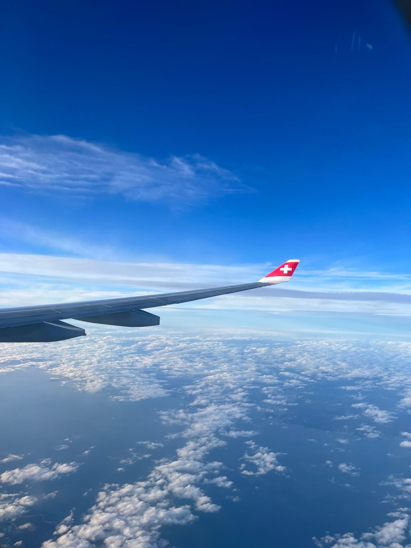 Airplane wing with Swiss flag in blue sky over clouds and ocean.