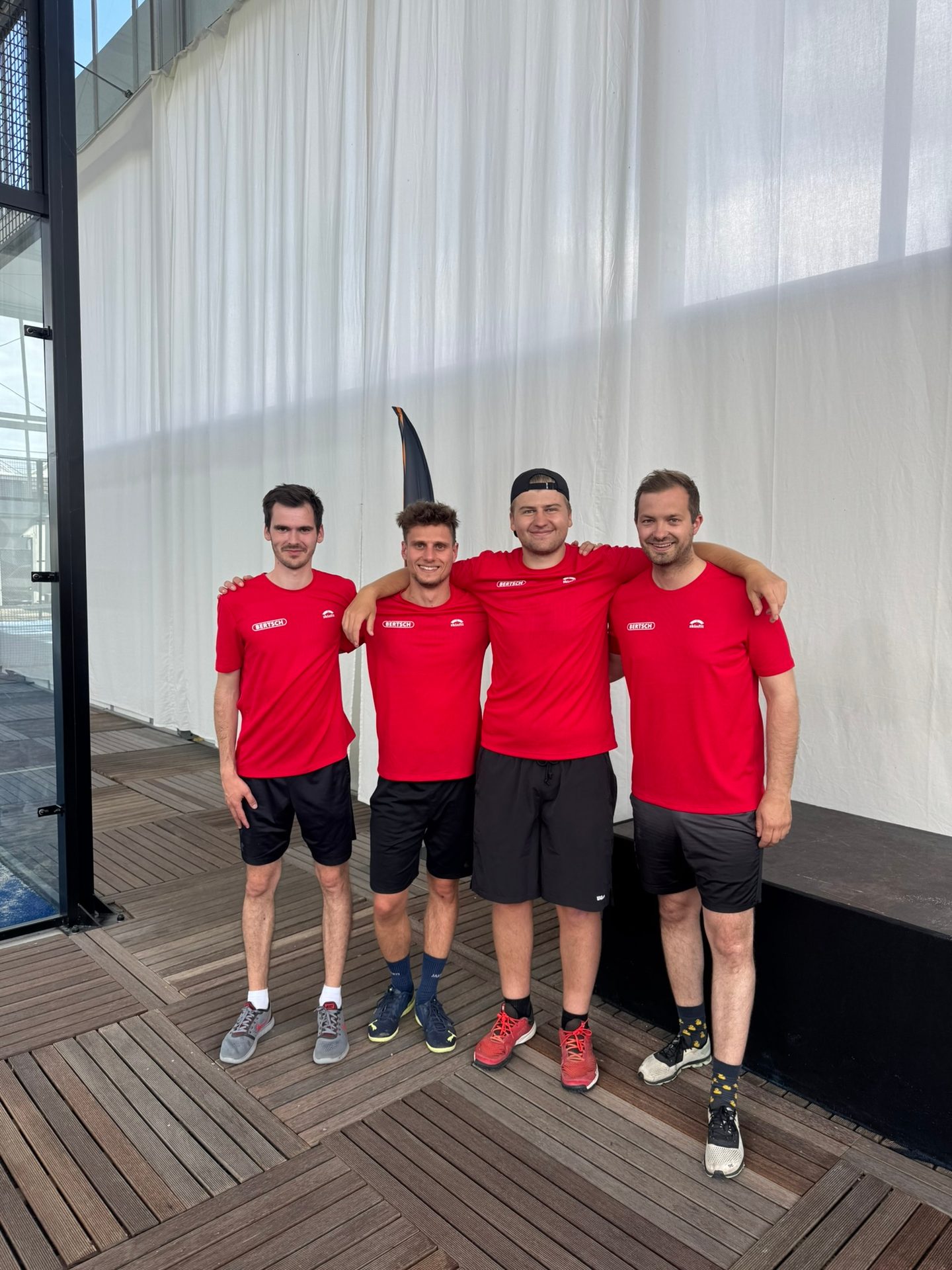 Four smiling men in red shirts and shorts on a deck.