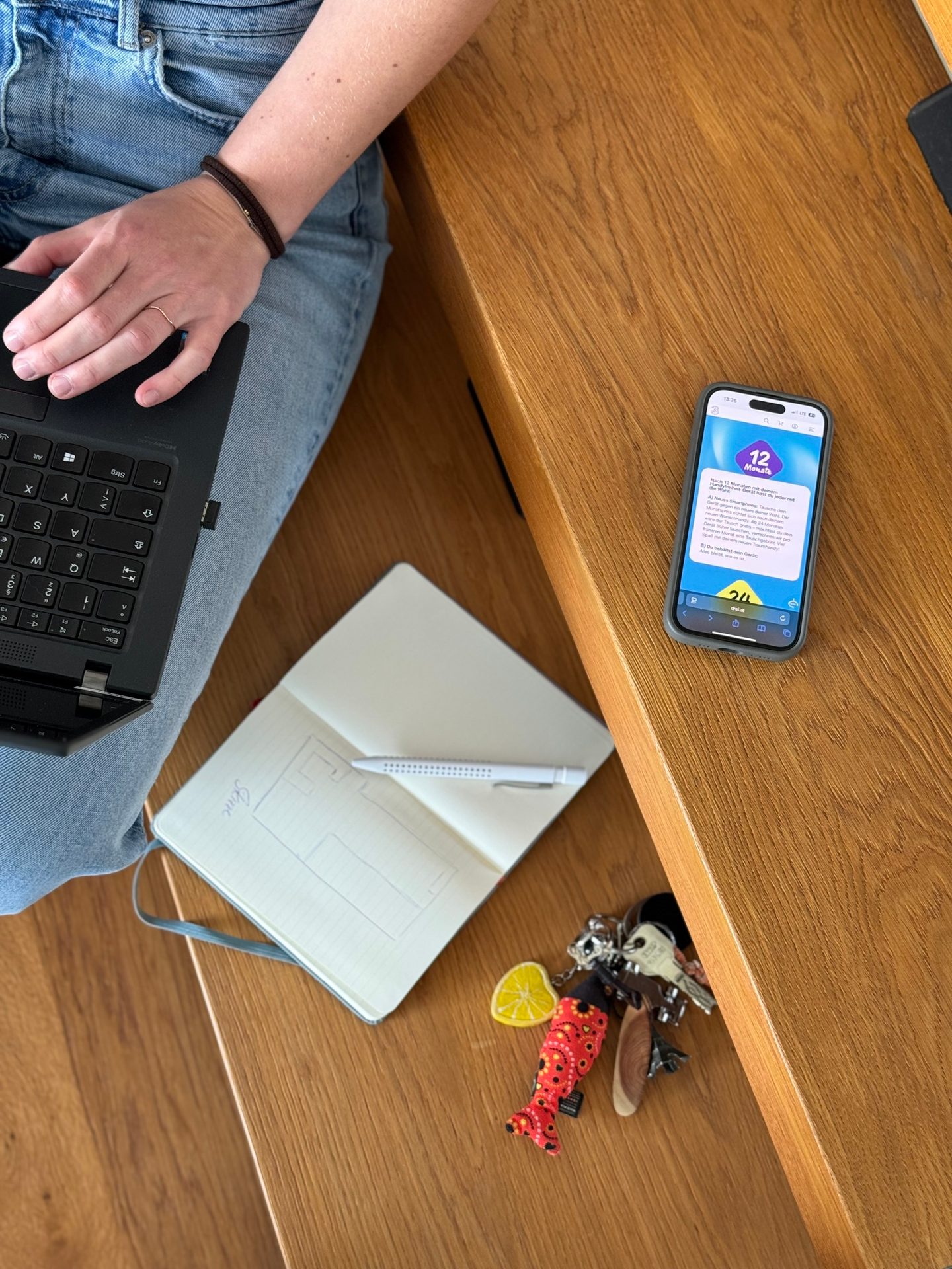 Overhead view: Hand on laptop, notebook, keys, and smartphone with app on wooden surface.