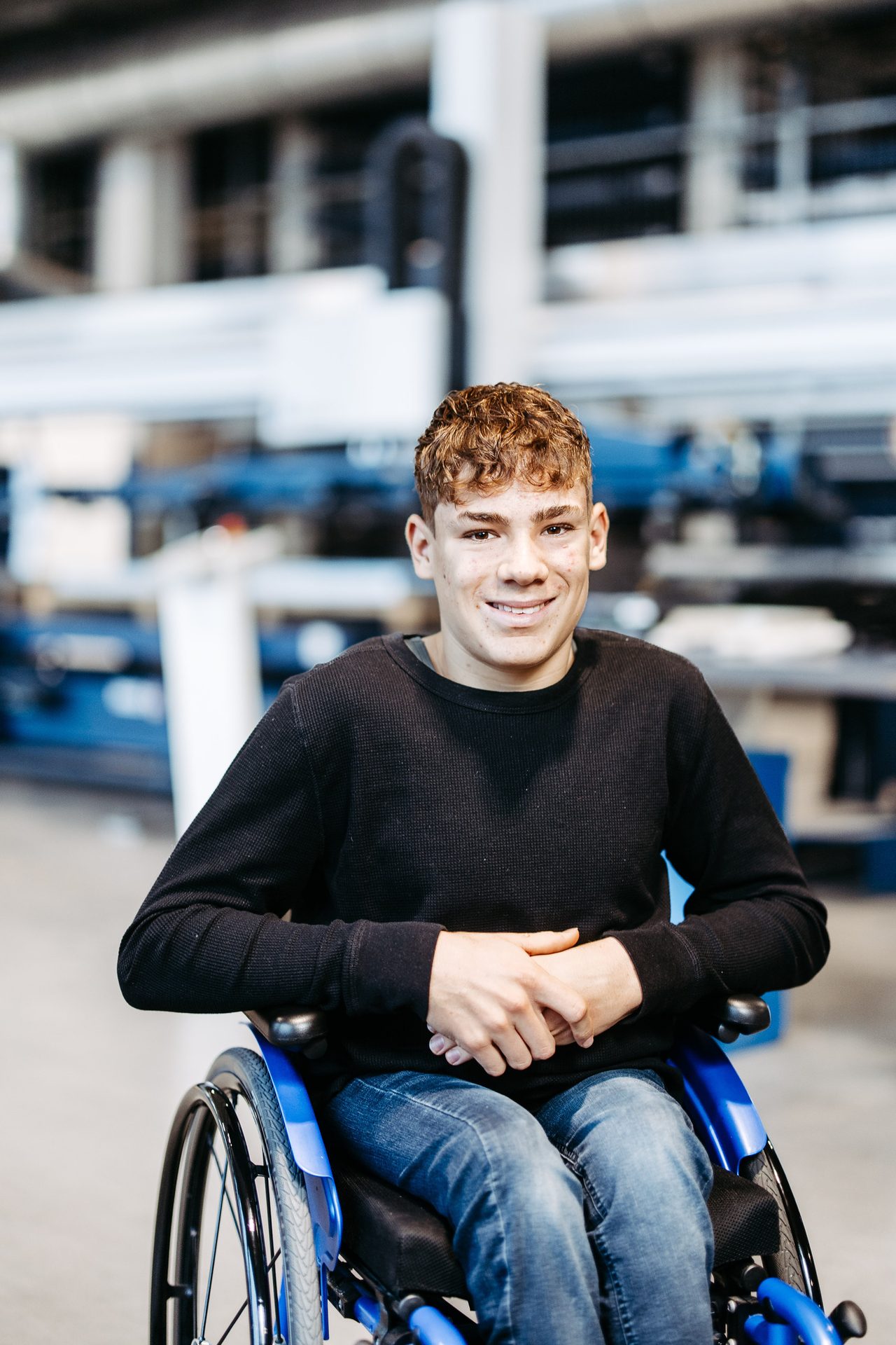 Young man with curly hair, sitting in a blue wheelchair and smiling at the camera in an industrial setting.