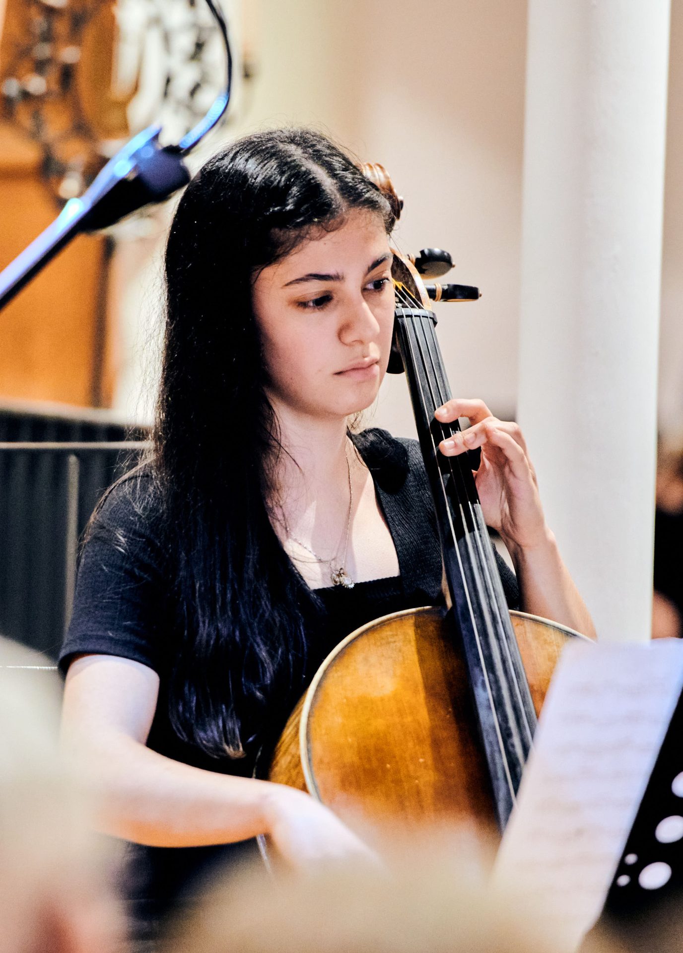Young woman playing cello intently, sheet music and microphone visible.