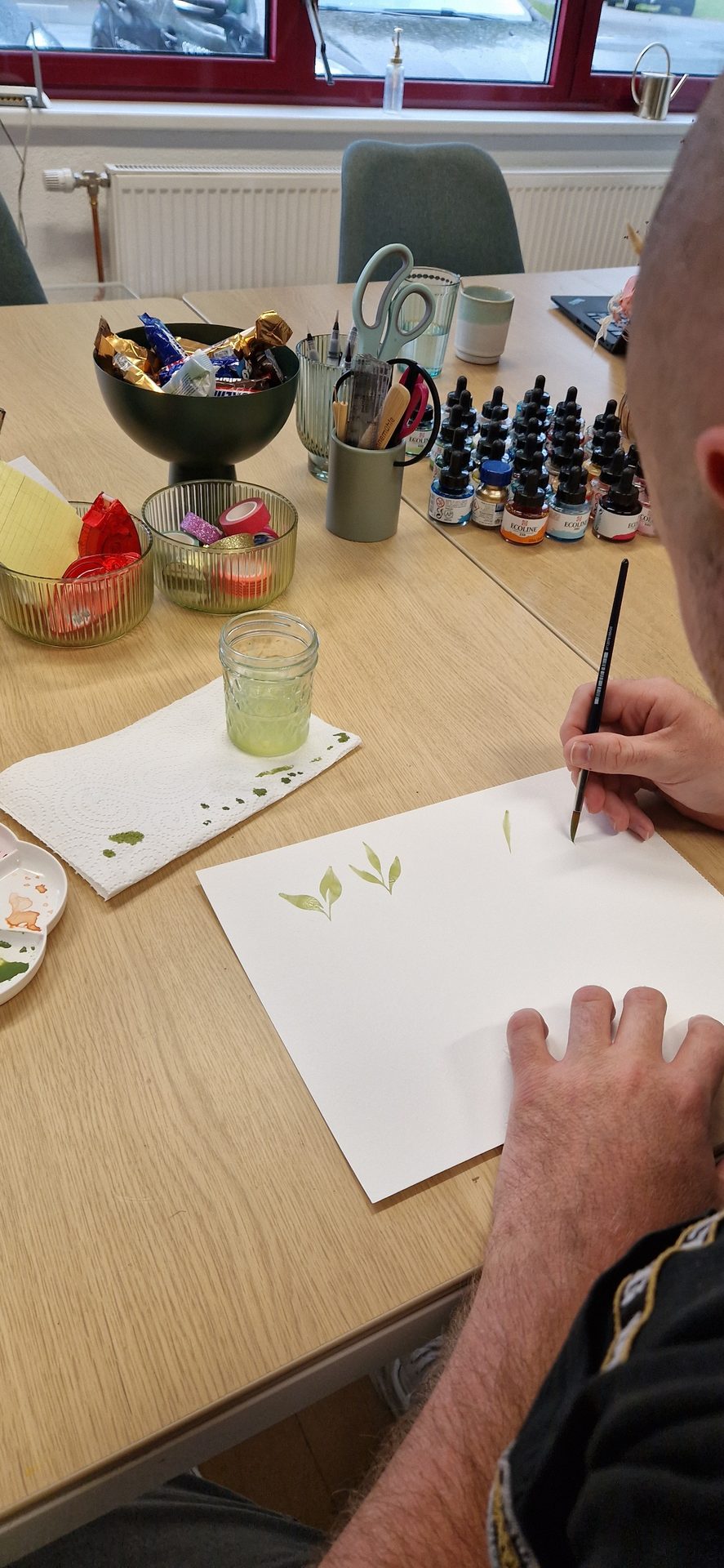 Person painting green leaves on white paper with a brush, surrounded by art supplies and candy.