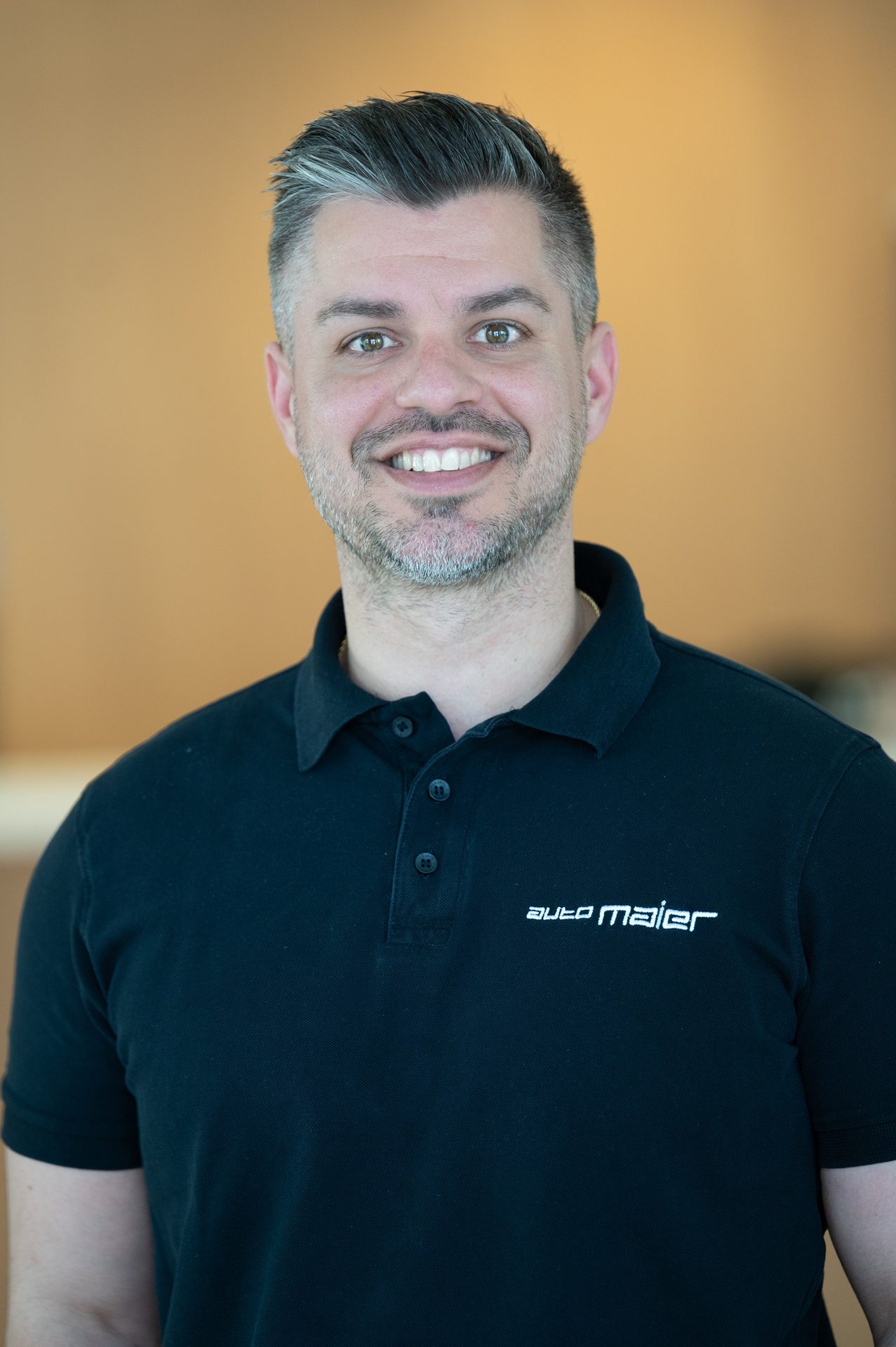 Smiling man with grey hair and beard, wearing a black "auto maier" polo shirt.