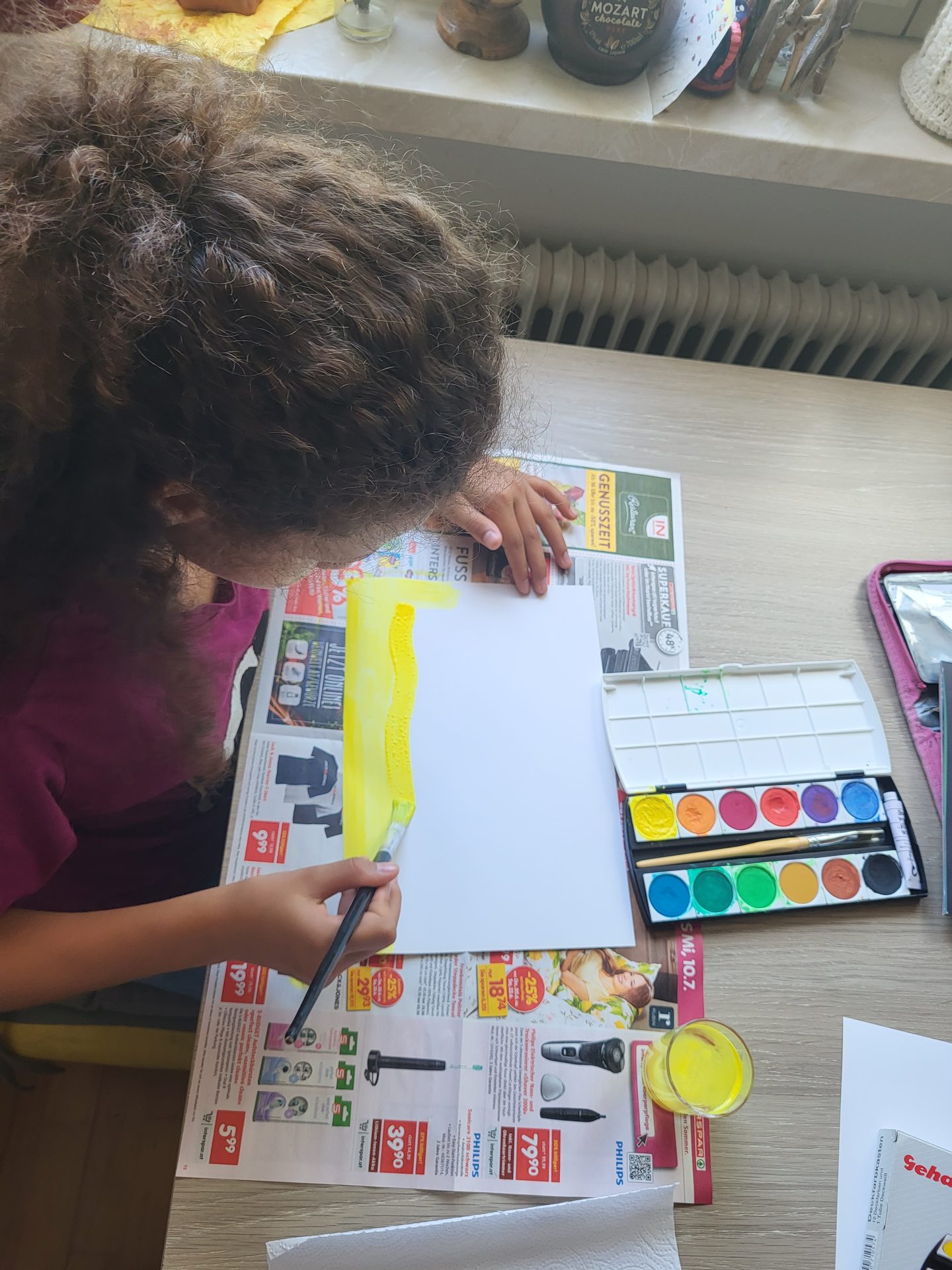 A child with curly hair painting a yellow strip with watercolors on paper, with a palette and water nearby.