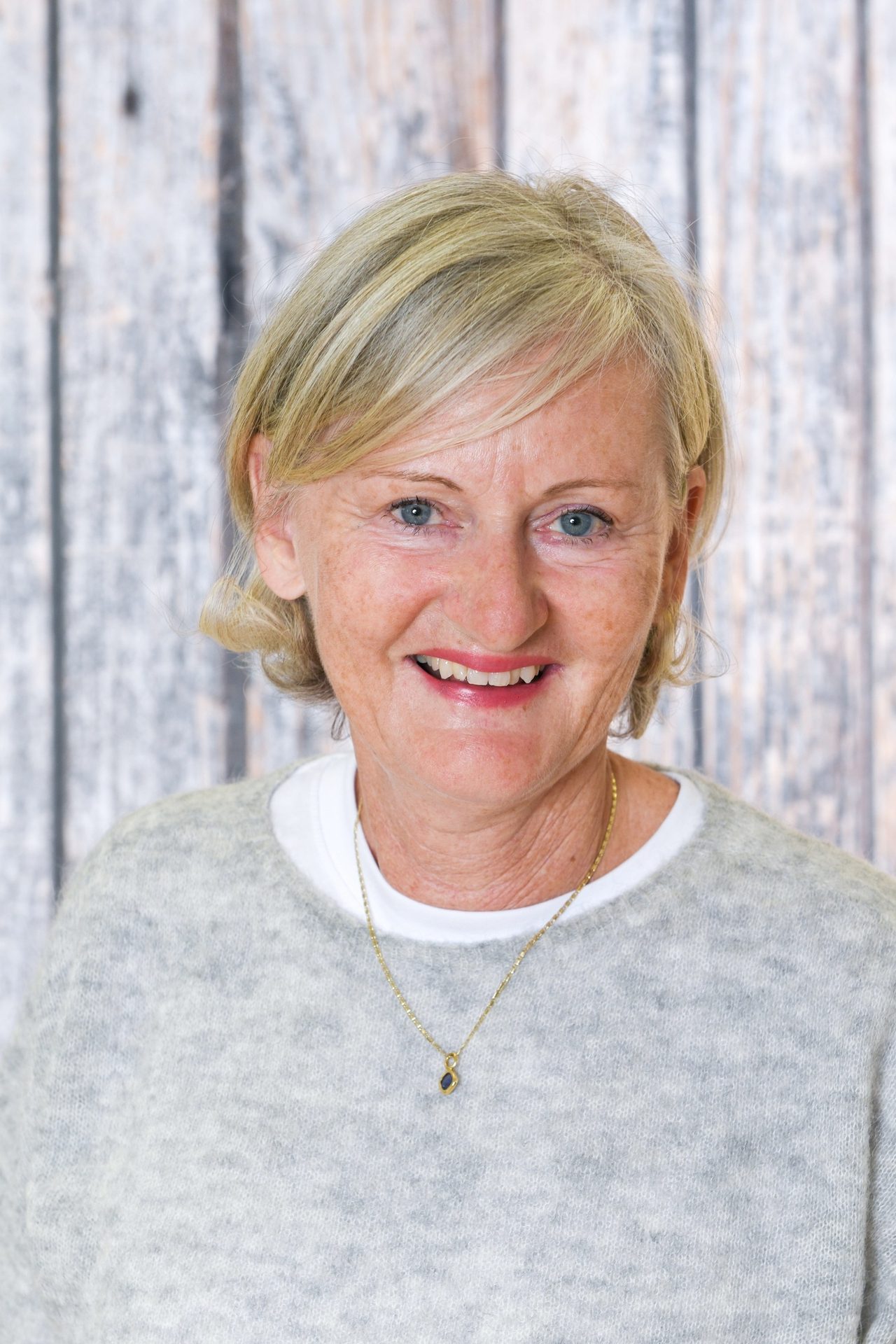 Smiling woman with blonde hair, blue eyes, wearing a gray sweater and gold necklace.