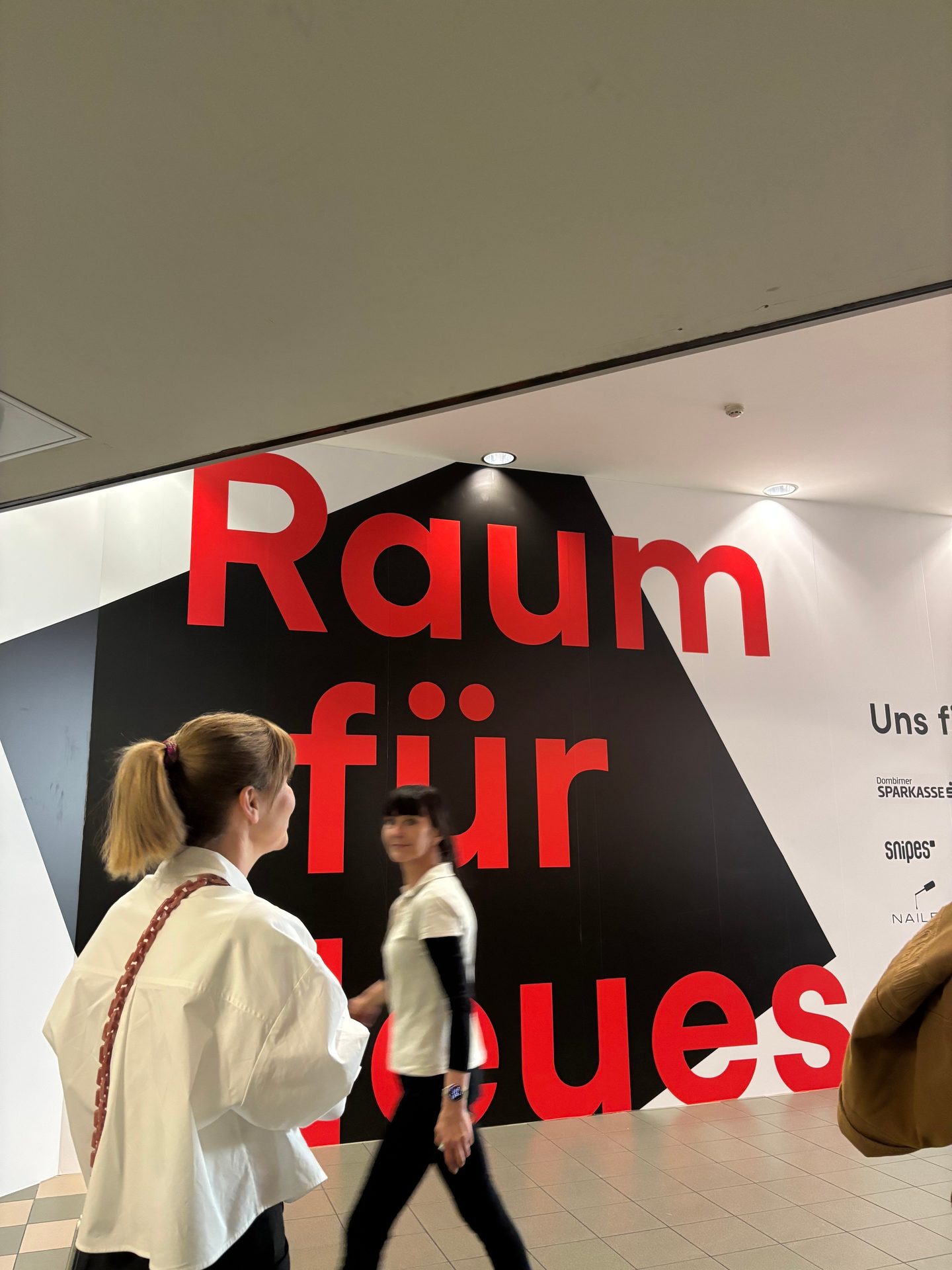 Two women walk past a large red sign reading "Raum für Neues" (Space for New Things) in a public space.