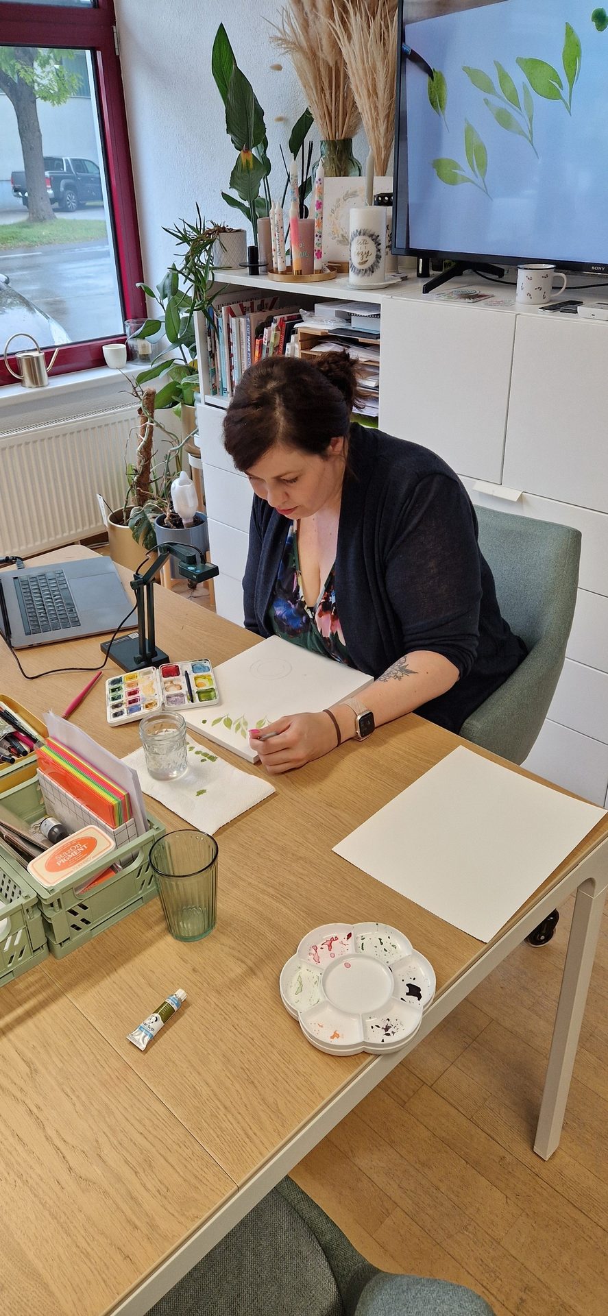 Woman painting watercolor leaves at a desk with a TV displaying leaf art.