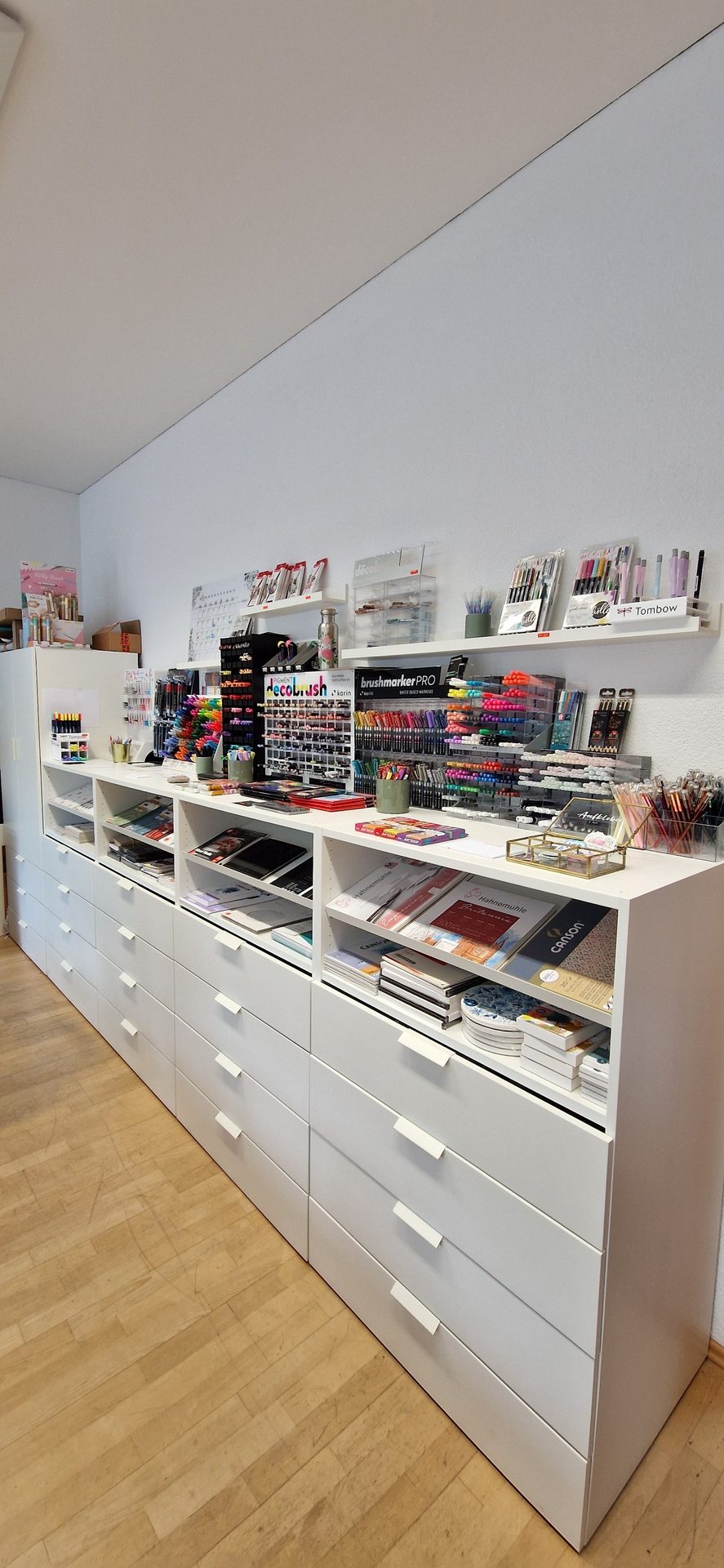 Long white art supply display unit with drawers, shelves of markers, pens, and paper, on wooden floor.