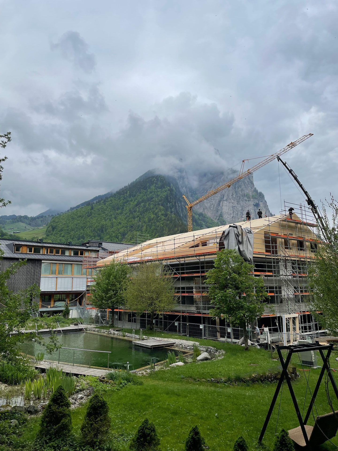 Building under construction with cranes, workers, a natural pool, and green mountains on a cloudy day.