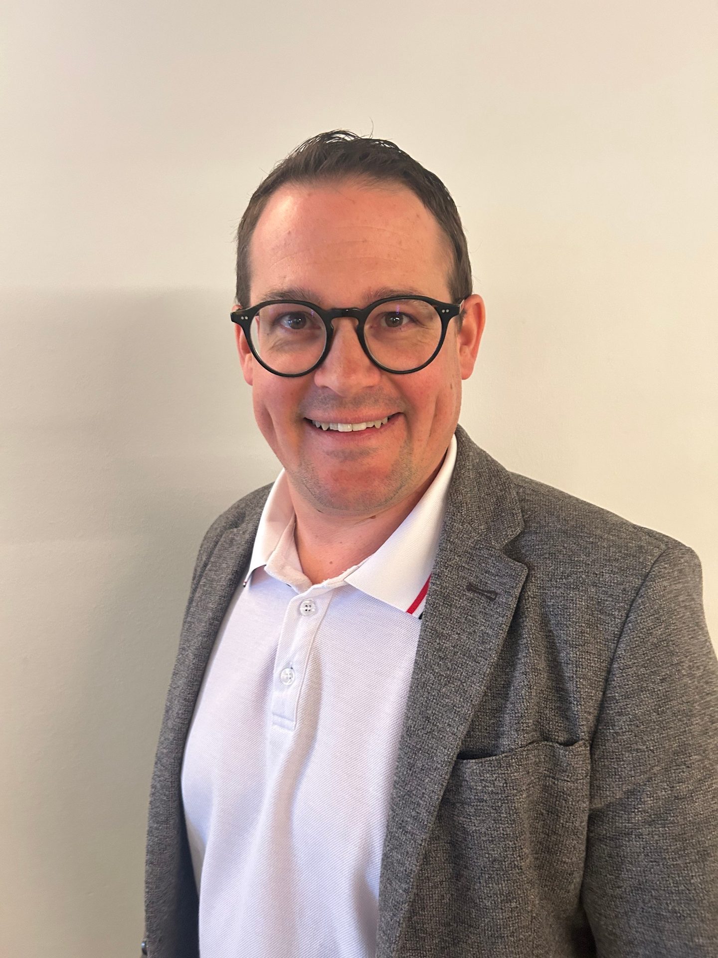 A smiling man wearing glasses, a white polo shirt, and a grey blazer, looking directly at the camera.
