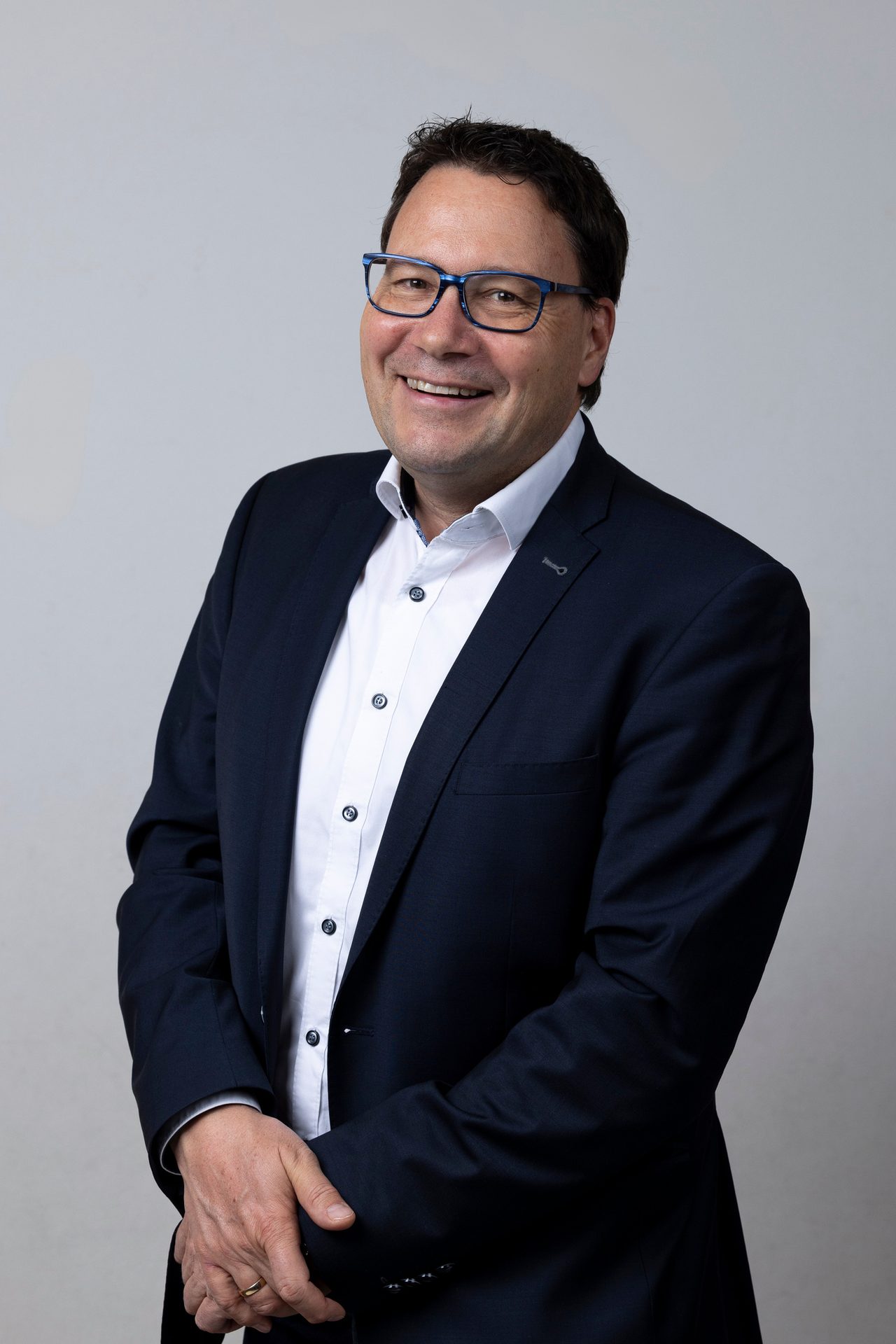 Smiling man in a dark suit and blue glasses, hands clasped.