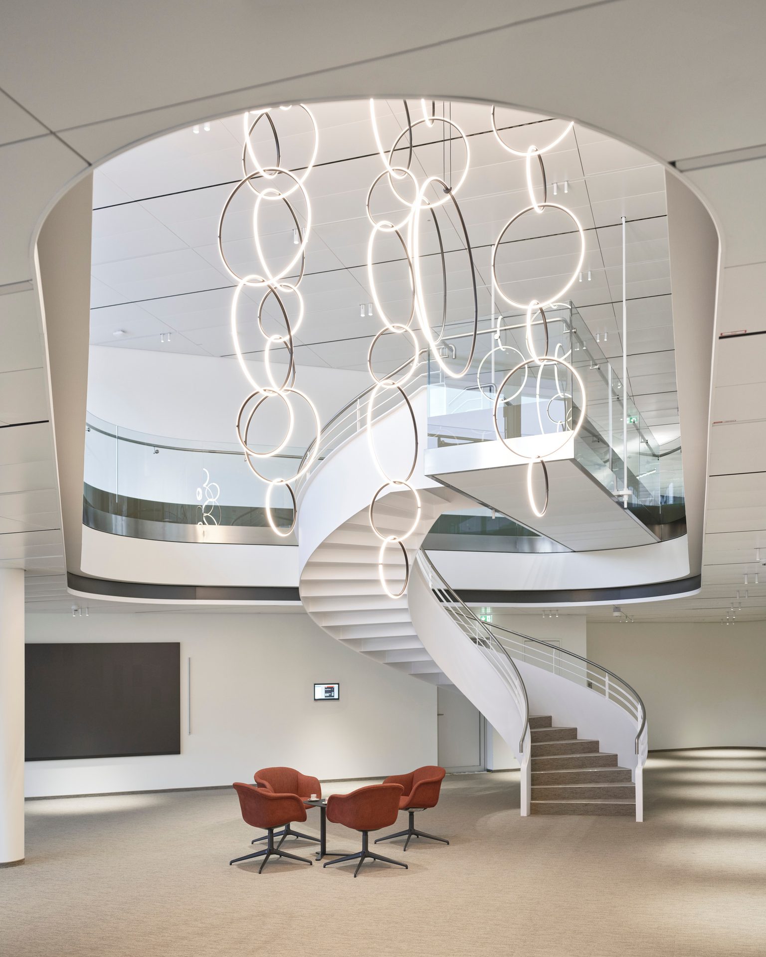 A modern lobby with a grand spiral staircase, a glowing ring chandelier, and a small seating area.