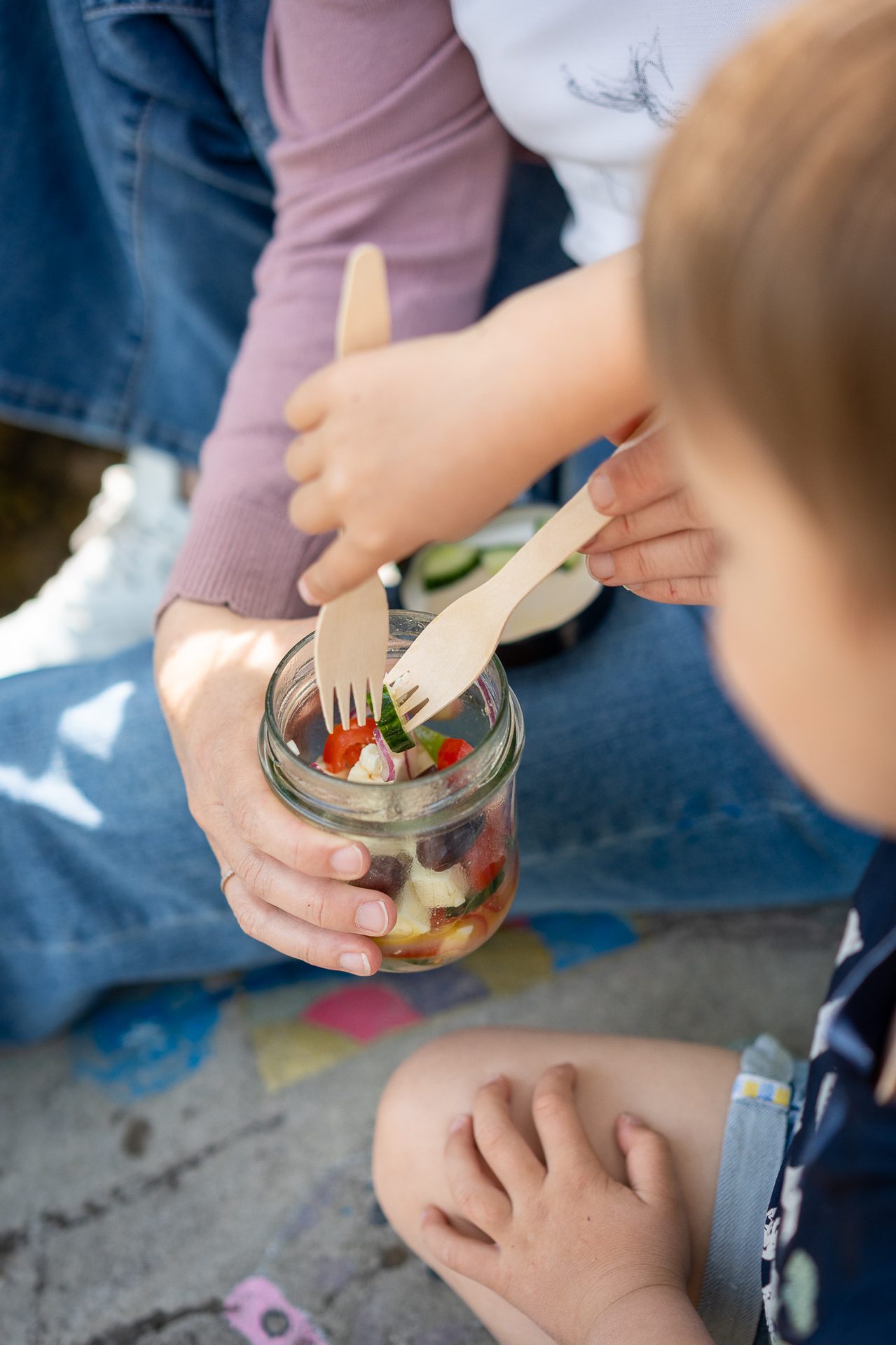 Close-up of an adult and child sharing a fresh vegetable and feta salad from a glass jar with wooden forks.