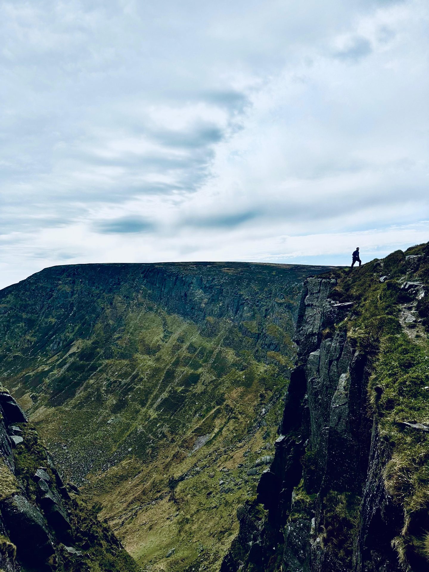 A person on a steep mountain cliff overlooks a deep valley under a cloudy sky.