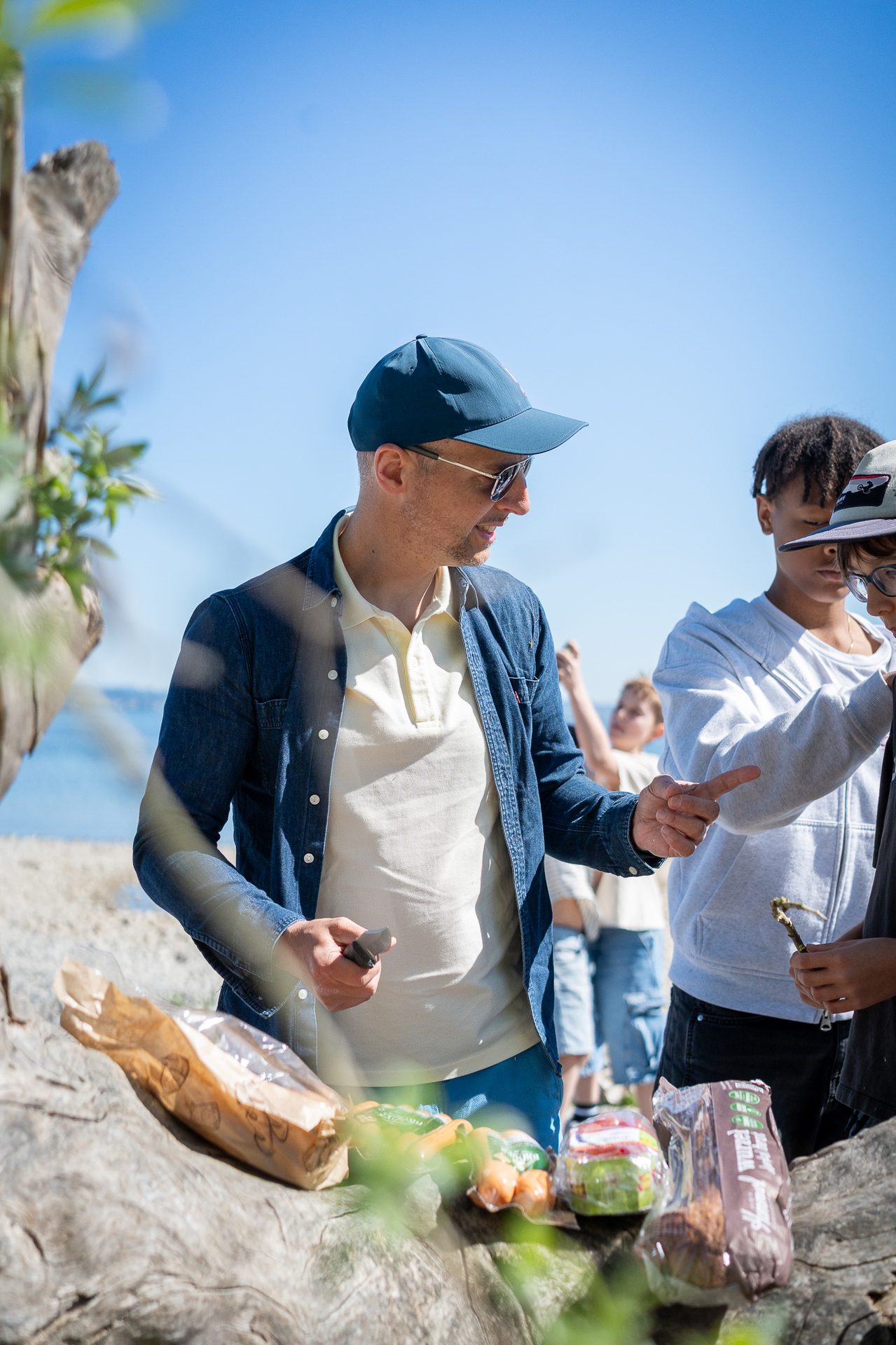 Man, cap, sunglasses, interacting with children by a log with food on a sunny day.