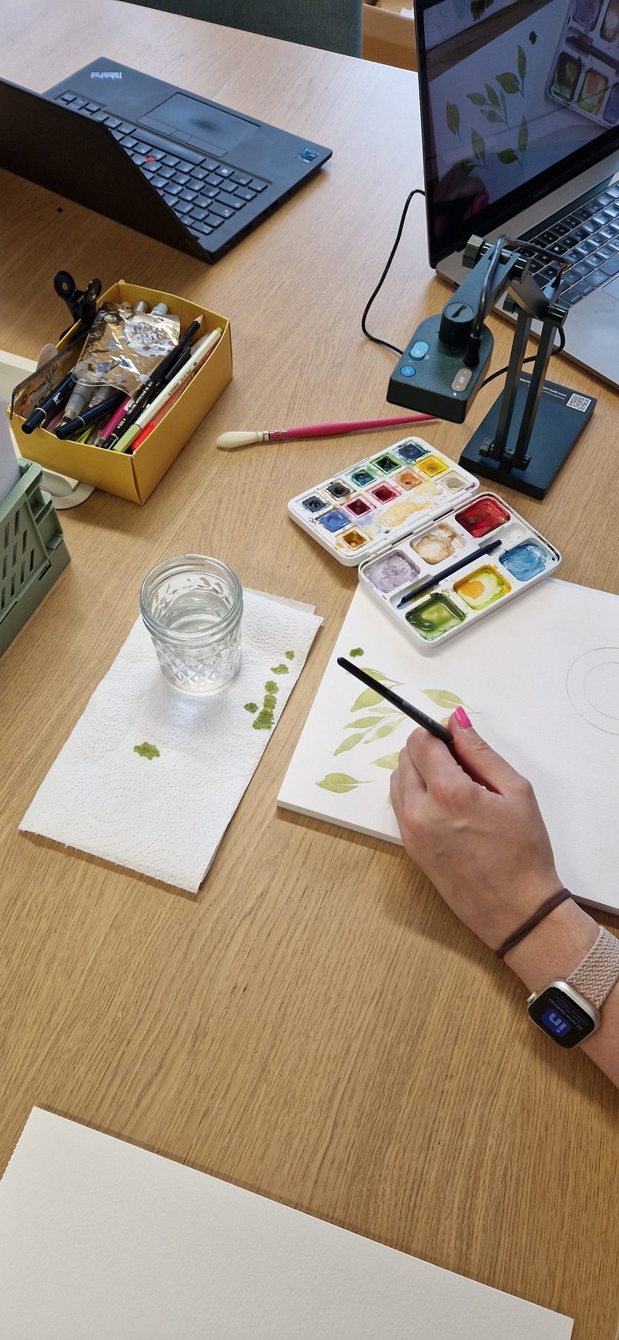 Person painting green leaves with watercolors at a desk.