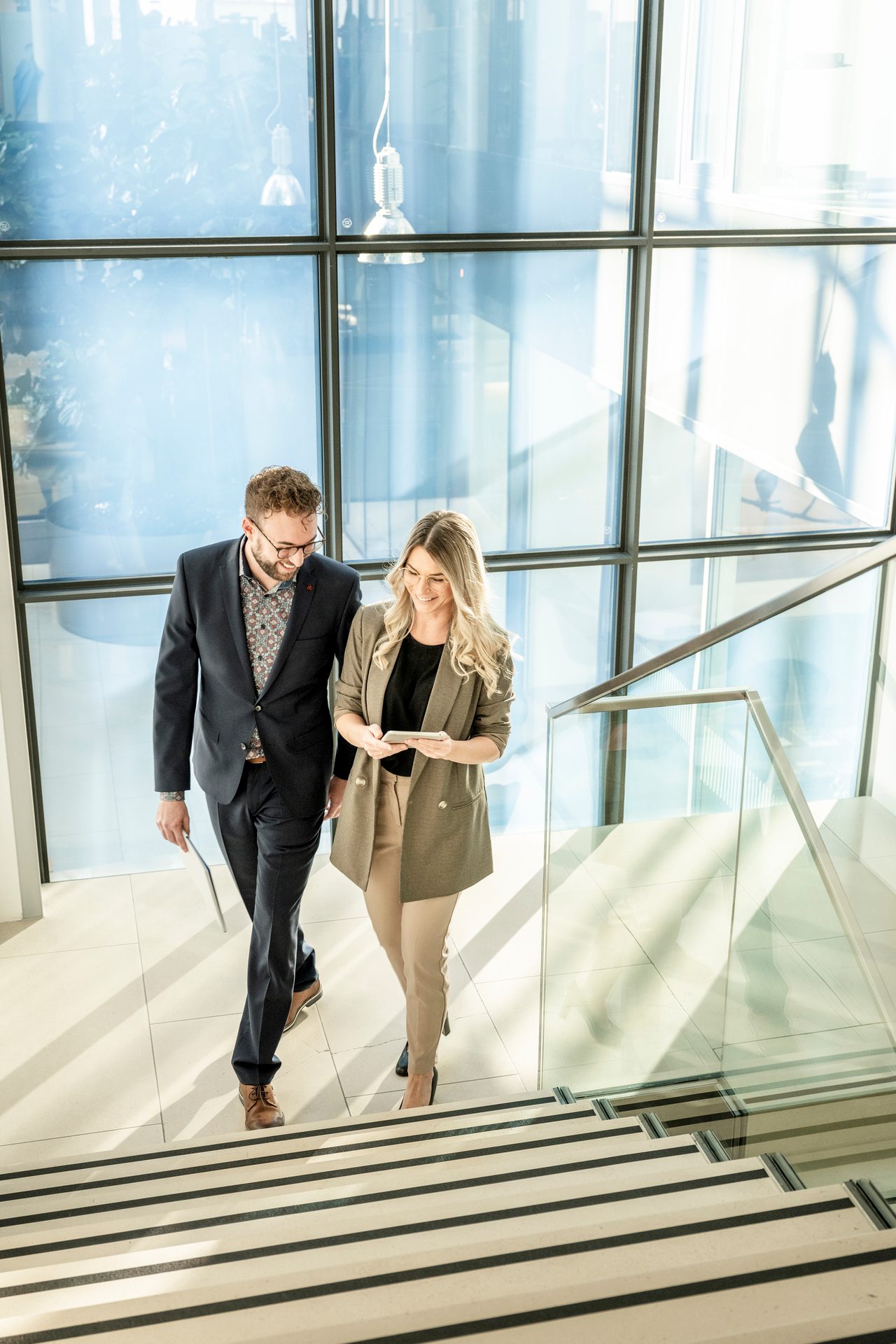 Two smiling business people walk up modern stairs, looking at devices, in a bright building.