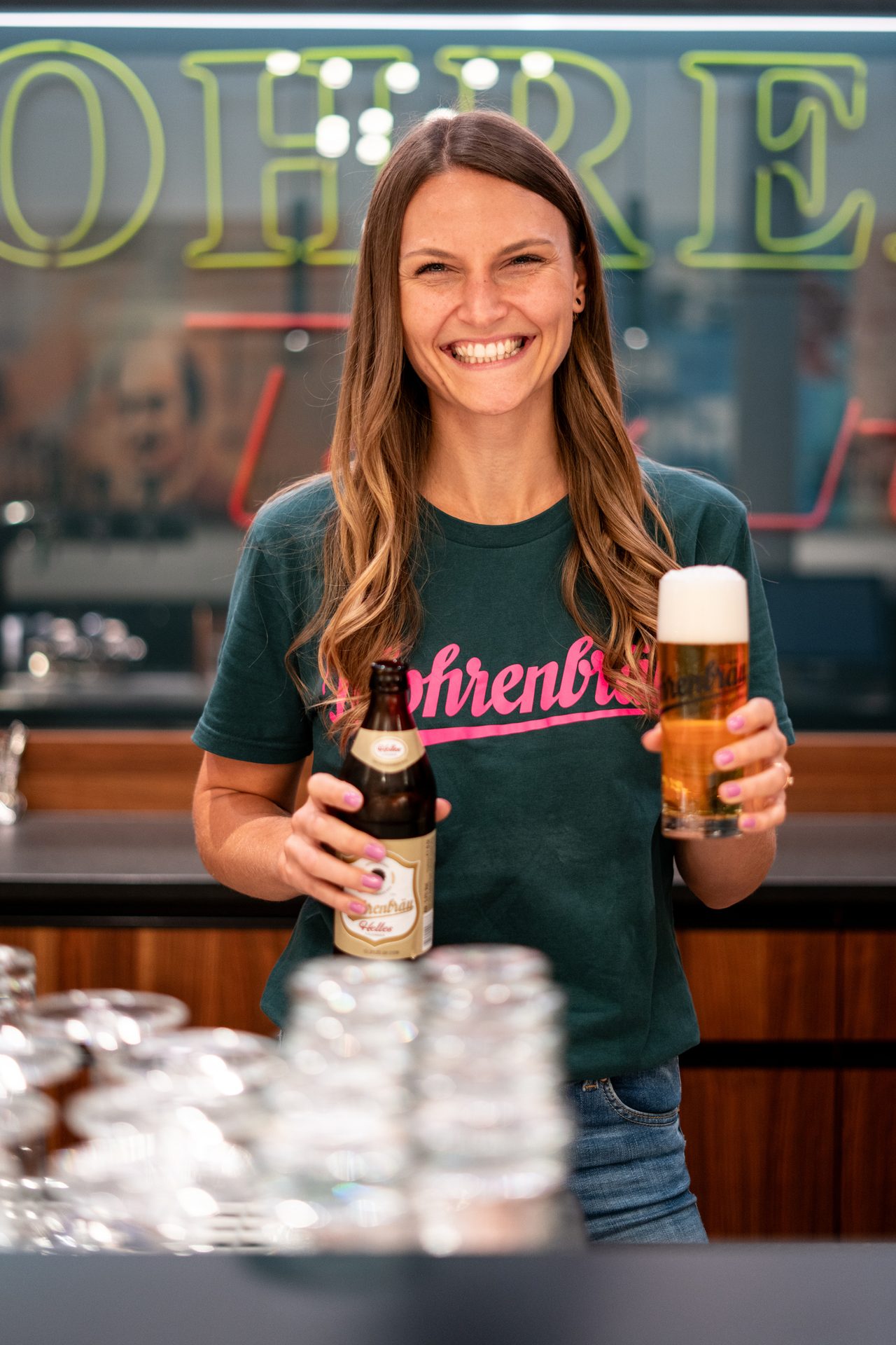 Smiling woman in a green t-shirt holds a beer bottle and a glass of beer.