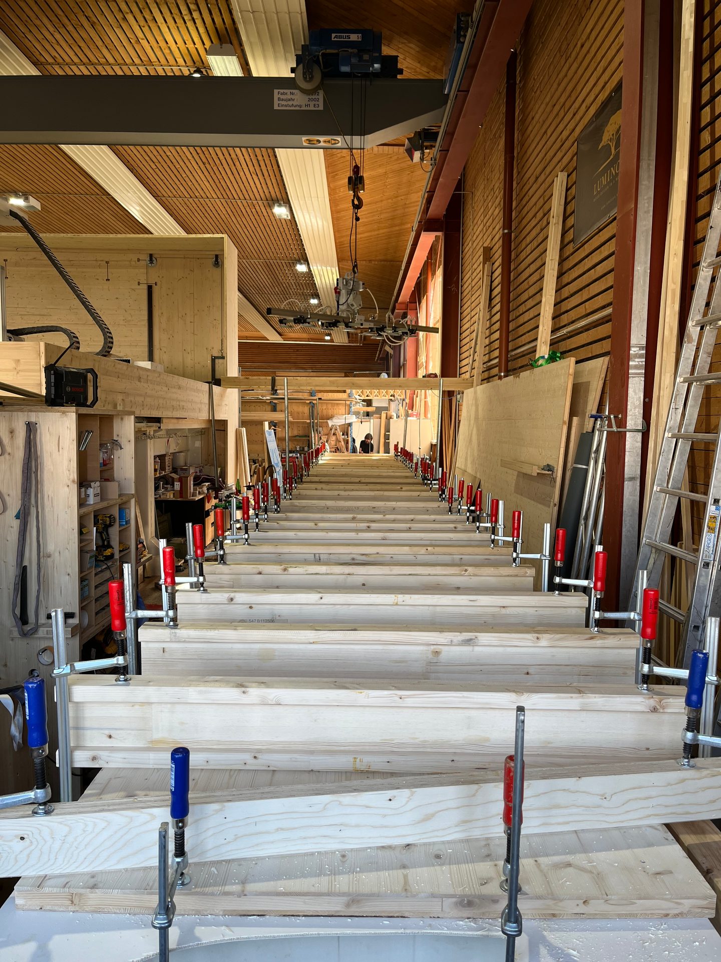 Long rows of wooden beams are clamped together in a spacious workshop with an overhead crane and various tools.