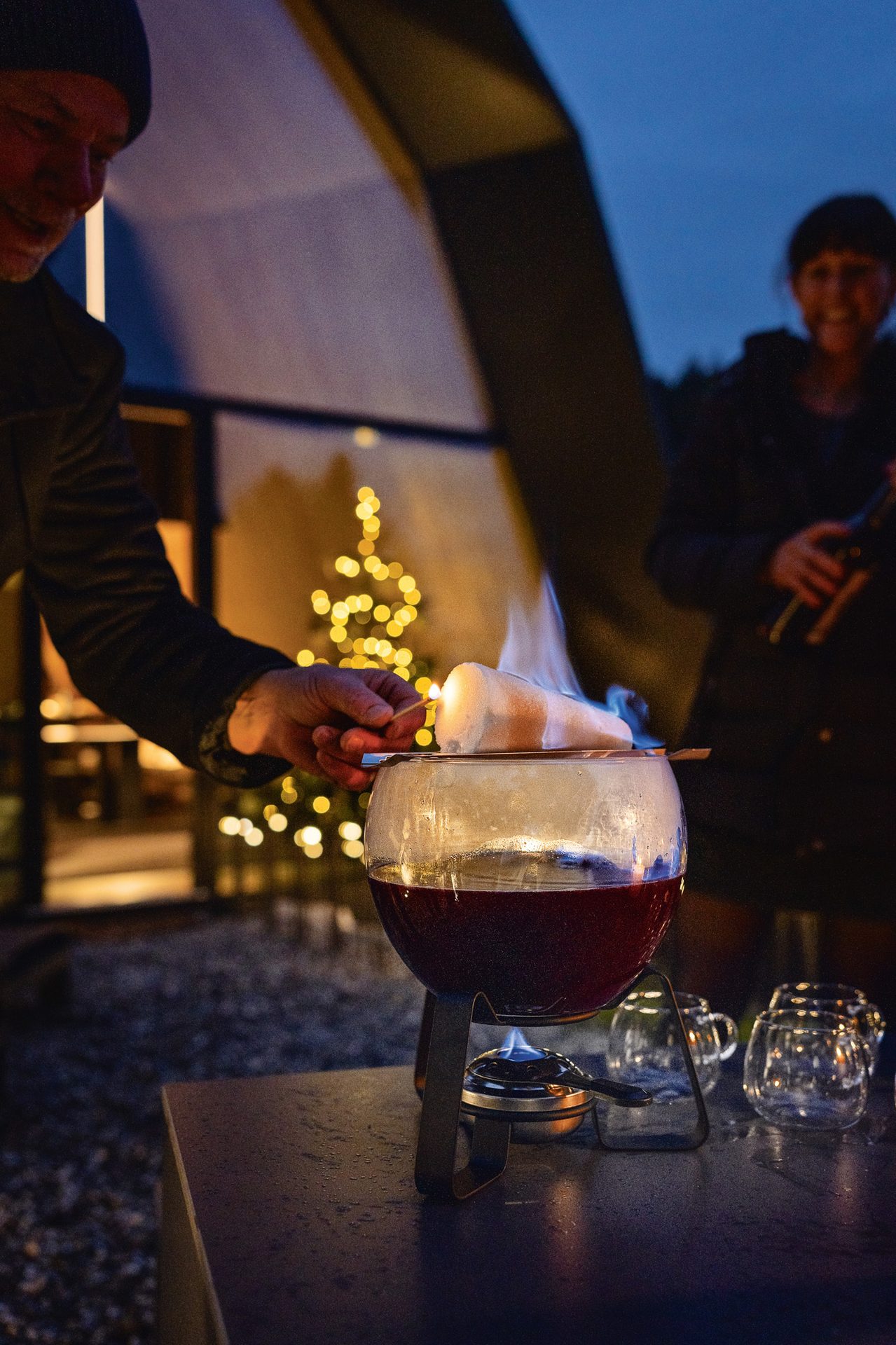 Man lights sugar for flaming punch, festive lights and a woman in background.