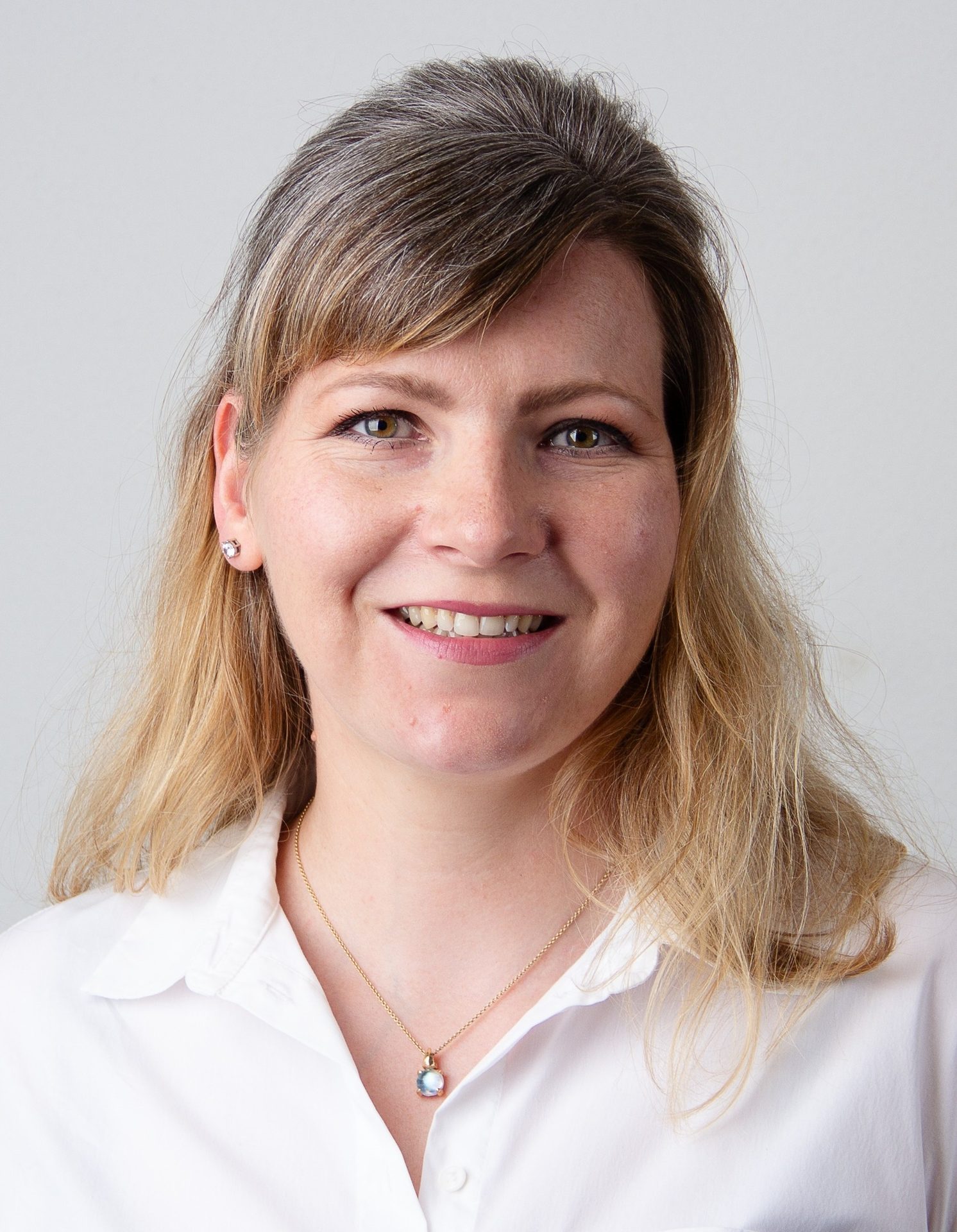 Headshot of a smiling woman with blonde hair, green eyes, wearing a white shirt and a necklace.