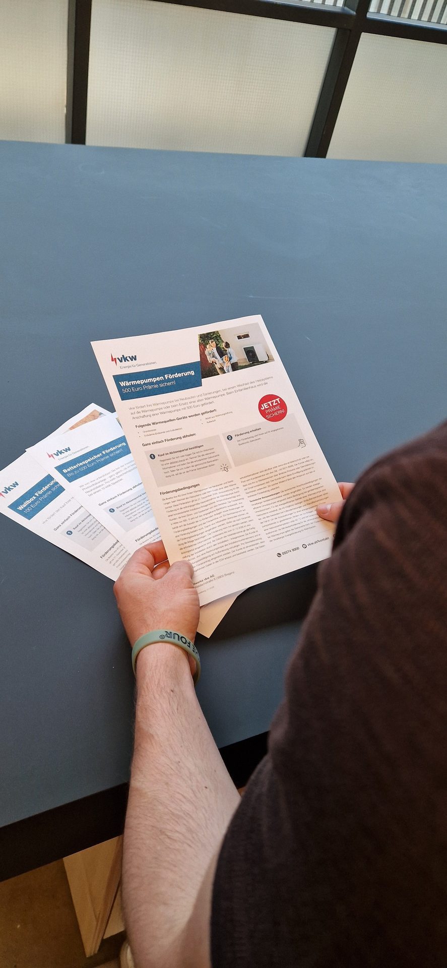 A person holds and reads a pamphlet on a table, titled 'Wärmepumpen Förderung 500 Euro Prämie sichern'.