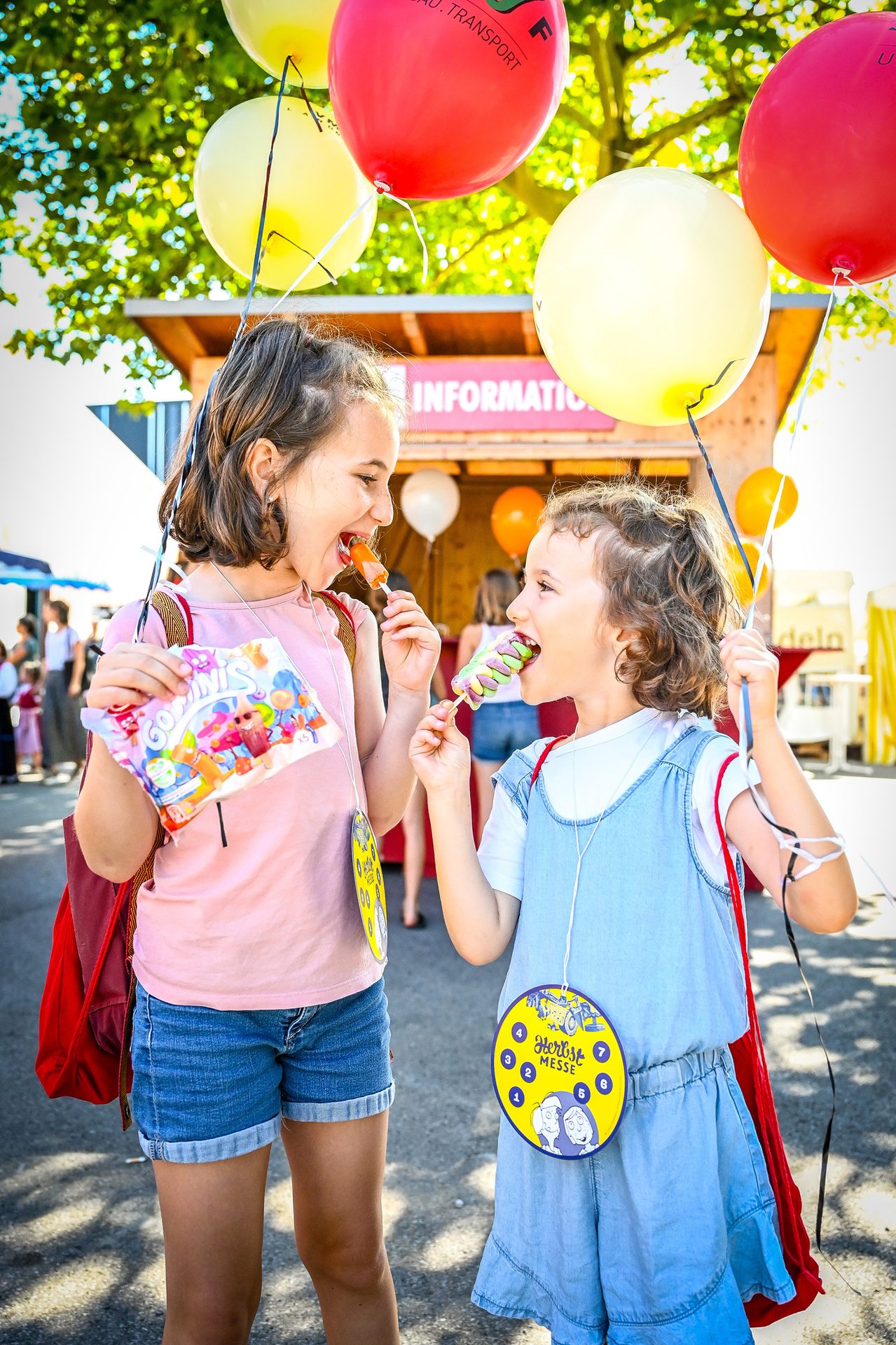 Two happy girls eating popsicles and holding balloons outdoors.