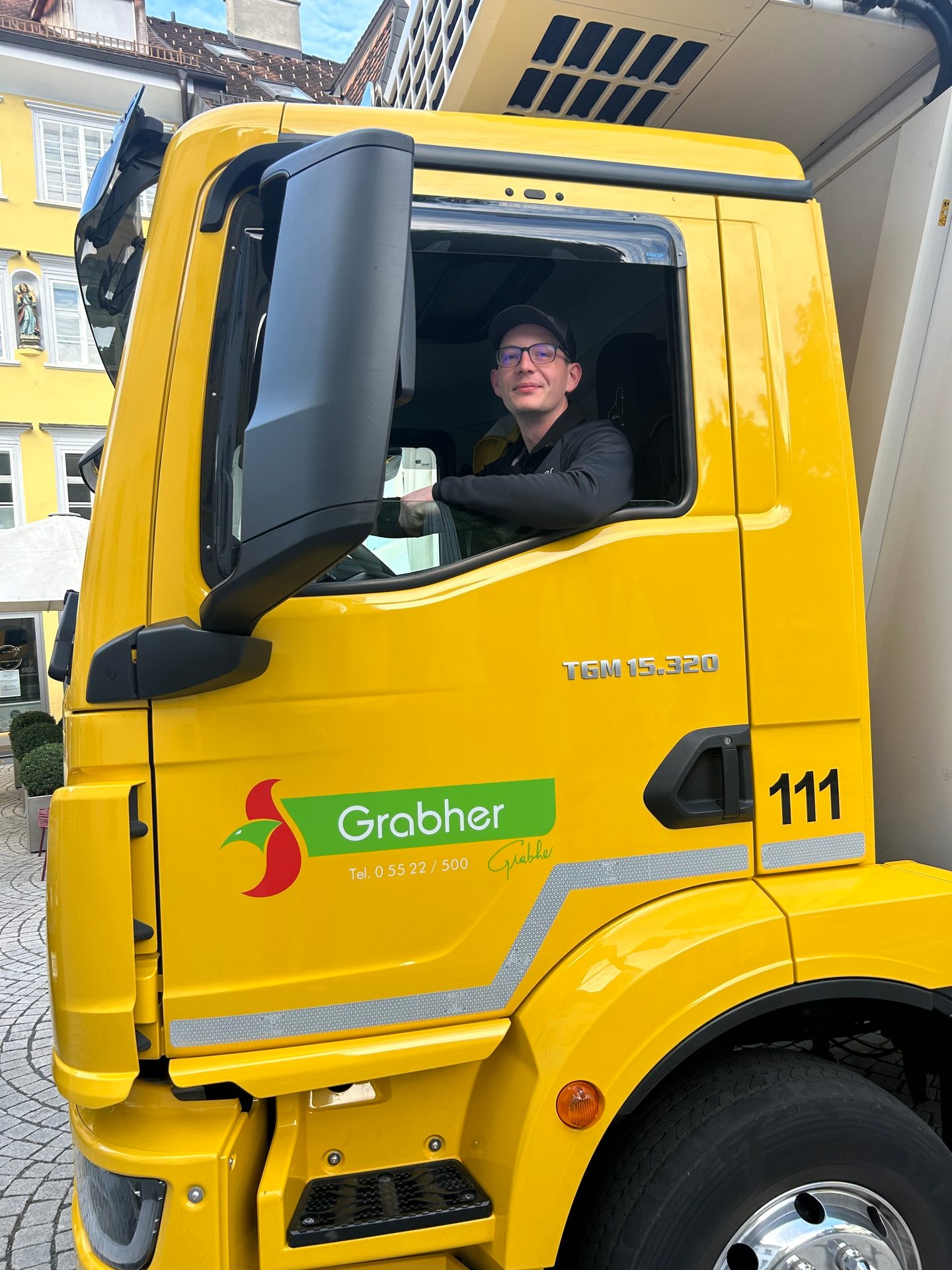 A man wearing glasses and a cap smiling from the driver's seat of a yellow Grabher truck.