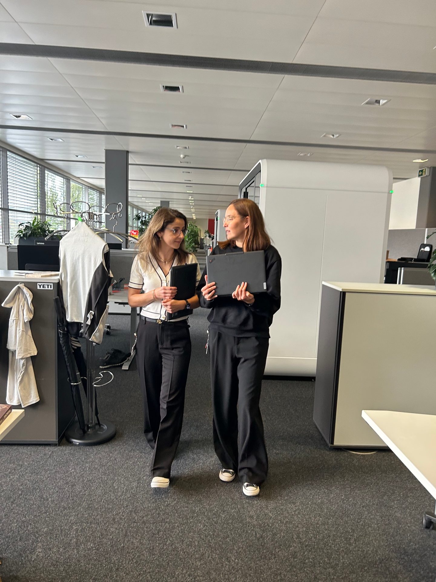 Two women walk and talk in a modern office, carrying laptops and a tablet.