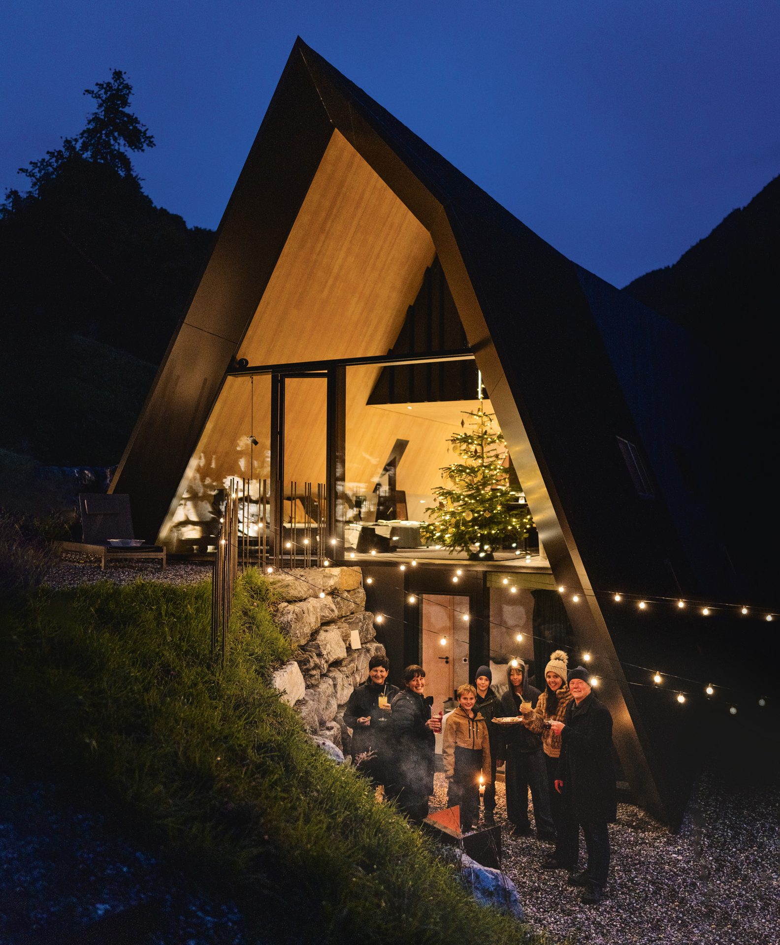A modern A-frame house at night with a Christmas tree inside, a group gathers outside by string lights.