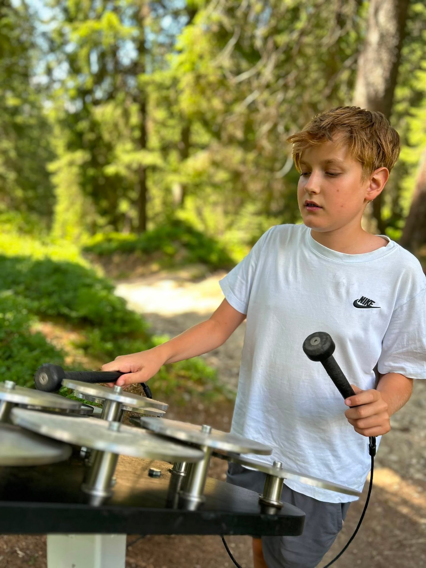 A young boy with blonde hair plays an outdoor xylophone-like instrument with mallets in a sunny forest.
