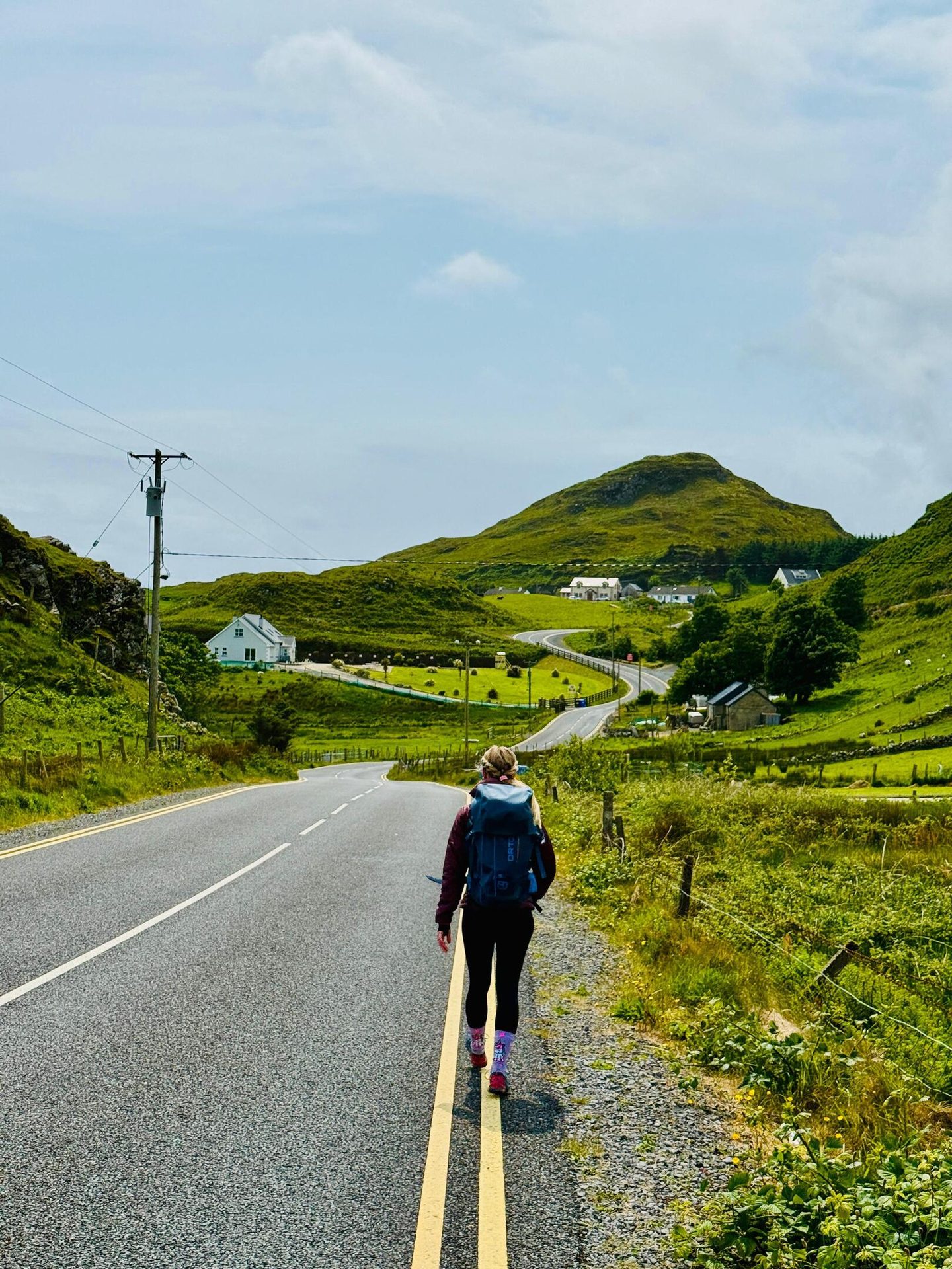 A hiker with a backpack walks on a winding road through green hills with distant houses under a cloudy sky.
