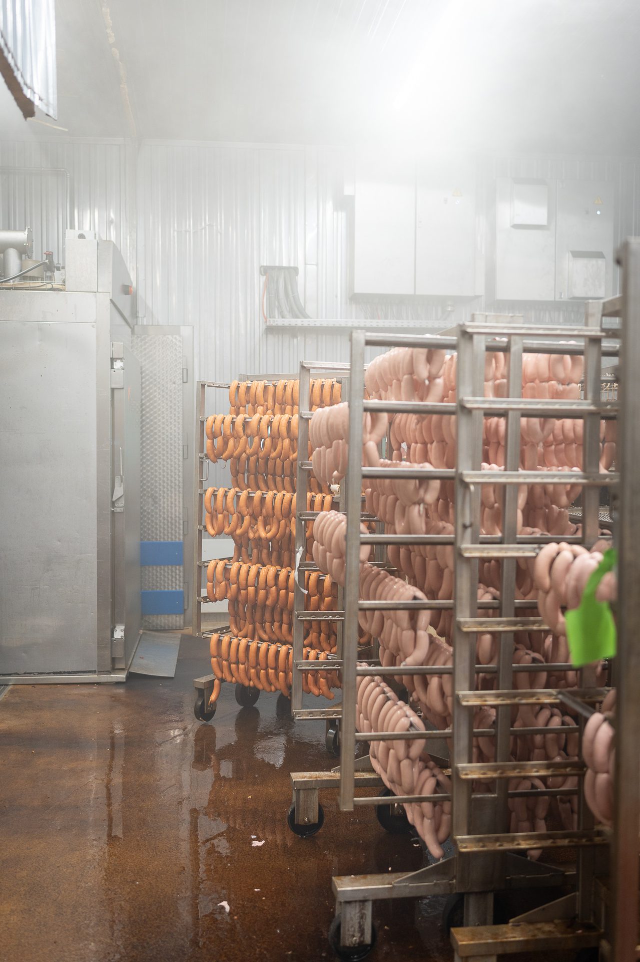 Racks of sausages, raw and processed, in a steamy meat processing plant with machinery.