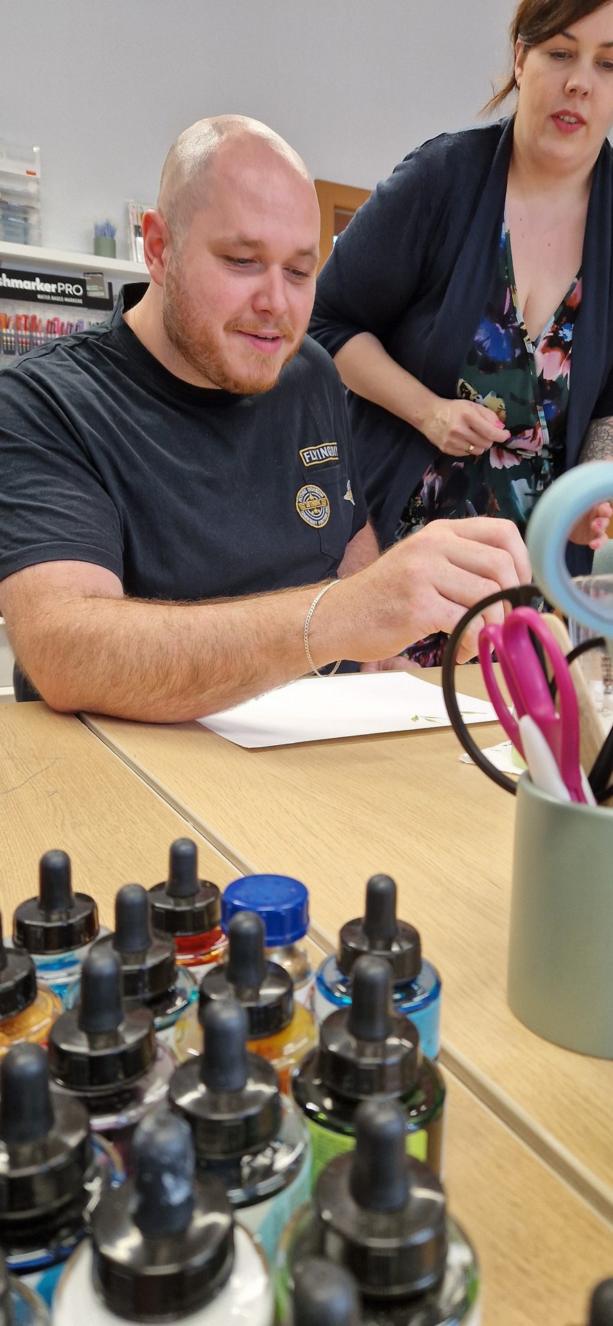 Man working with art supplies at table, woman observing.