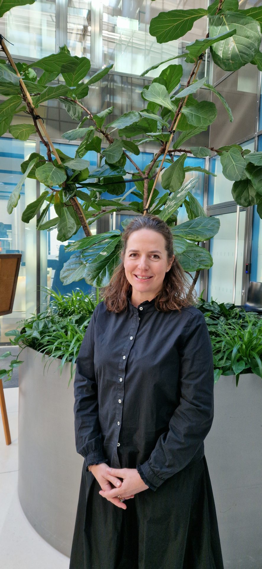Smiling woman in black shirt, hands clasped, by large plants & modern building.