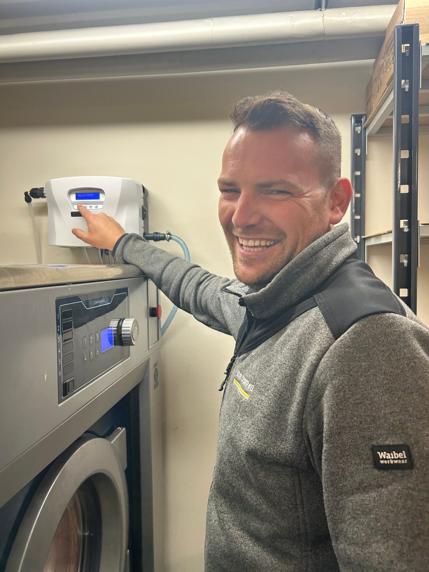 A smiling man in a grey workwear jacket points at a control panel of an industrial washing machine.