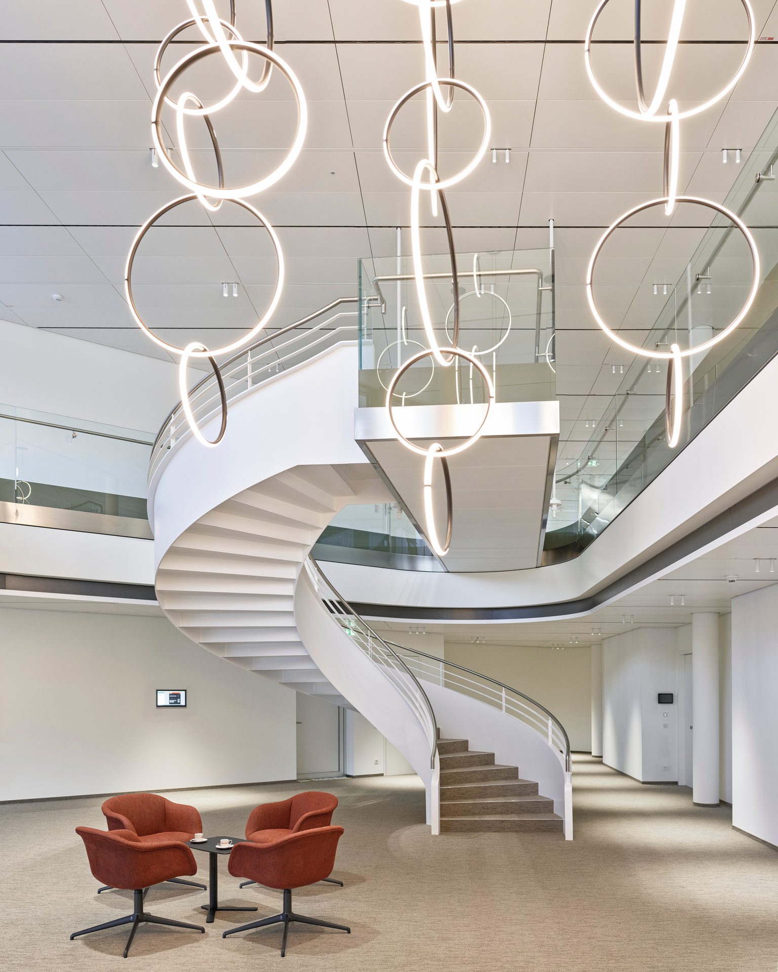 Modern lobby with a grand white spiral staircase, striking circular lights, and red armchairs.