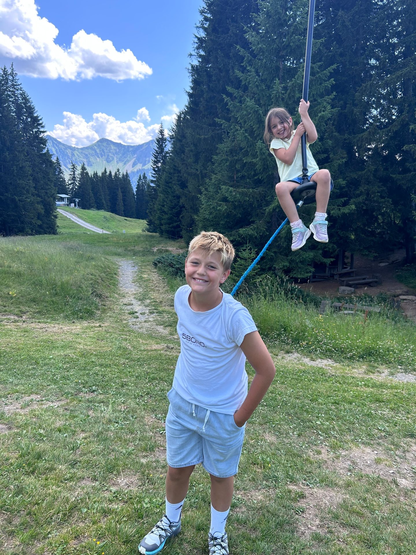 A boy smiling at the camera while a girl ziplines in the background with mountains and trees.