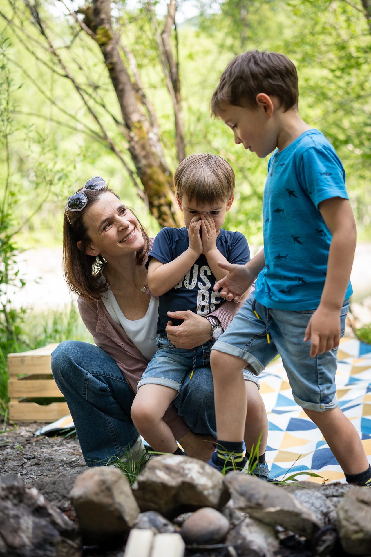 A smiling woman embraces a boy covering his face, while another boy stands nearby in a lush outdoor setting.