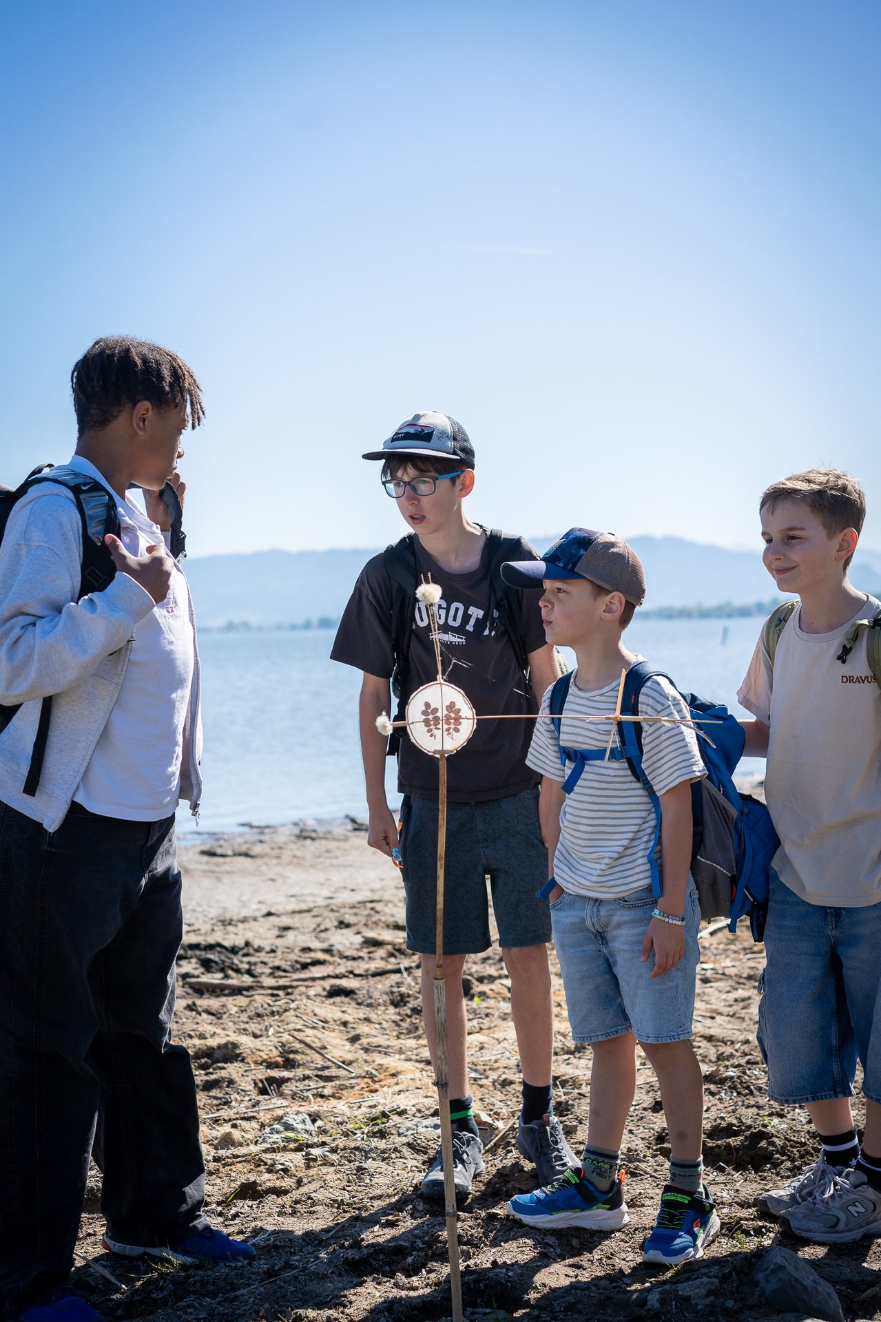 Four boys with backpacks on a sunny day by a lake, looking at something on the ground.